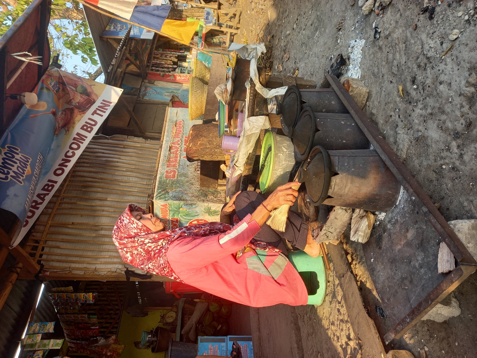 The image depicts an outdoor scene in Subang, Indonesia, focusing on a woman preparing food. The woman, wearing a red and white patterned hijab and a pink long-sleeved shirt, is seated on a small green stool. She is actively engaged in fanning flames beneath a cooking setup, which consists of three dark, cylindrical pots placed on a metal grill. The pots appear to be made of a heavy material, possibly cast iron or clay. Beside her, on a wooden table, are various cooking implements, including a purple mug, green plastic lids, and a white cloth.
The environment suggests a humble roadside food stall. In the background, a corrugated metal structure serves as a stall, adorned with banners. One prominent banner reads "LEMON MADU" and another mentions "SURABI ONCOM BU TINI," indicating the type of food sold. Shelves behind the woman hold numerous small packets of goods, likely snacks or ingredients. To the left, stacked blue boxes and some green coconuts are visible. The ground is uneven and dusty, with scattered debris and evidence of previous fires, such as ash and charcoal. The lighting suggests it is daytime, with sunlight casting shadows. The overall impression is one of everyday life and small-scale commerce in an Indonesian setting.