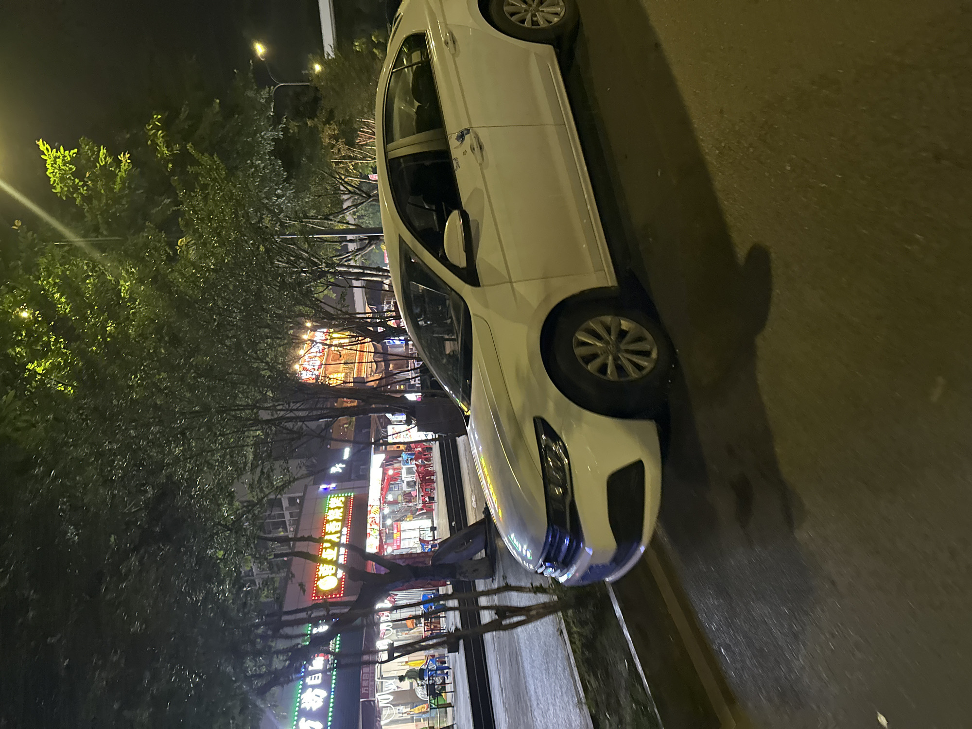 The scene depicts a nighttime urban street in the Chongqing High-tech Zone, China. A white four-door sedan is parked curb-side on the asphalt pavement in the foreground, oriented slightly towards the viewer's left. The vehicle's driver's side displays a small blue and white graphic on the front door.
To the left of the car, a raised sidewalk is lined with numerous deciduous trees featuring dense green foliage, partially obscuring the storefronts behind them. Beyond the trees, multiple commercial establishments are visible, illuminated by vibrant neon signs displaying Chinese characters. One prominent sign, largely red and yellow, reads "芭亚入巷烧烤" (Baya Ruxiang BBQ), while another on the far left exhibits green and white lighting.
Outdoor seating arrangements, including tables and red chairs, are set up in front of several establishments, indicating dining or gathering areas. Multiple individuals are present in these areas; one person in a light-colored shirt stands near tables on the left, while others are seated or standing further back.
Overhead streetlights emit a warm yellow glow, illuminating parts of the street and foliage. The asphalt pavement in the foreground shows some reflective patches. The sky is dark, consistent with nighttime conditions.