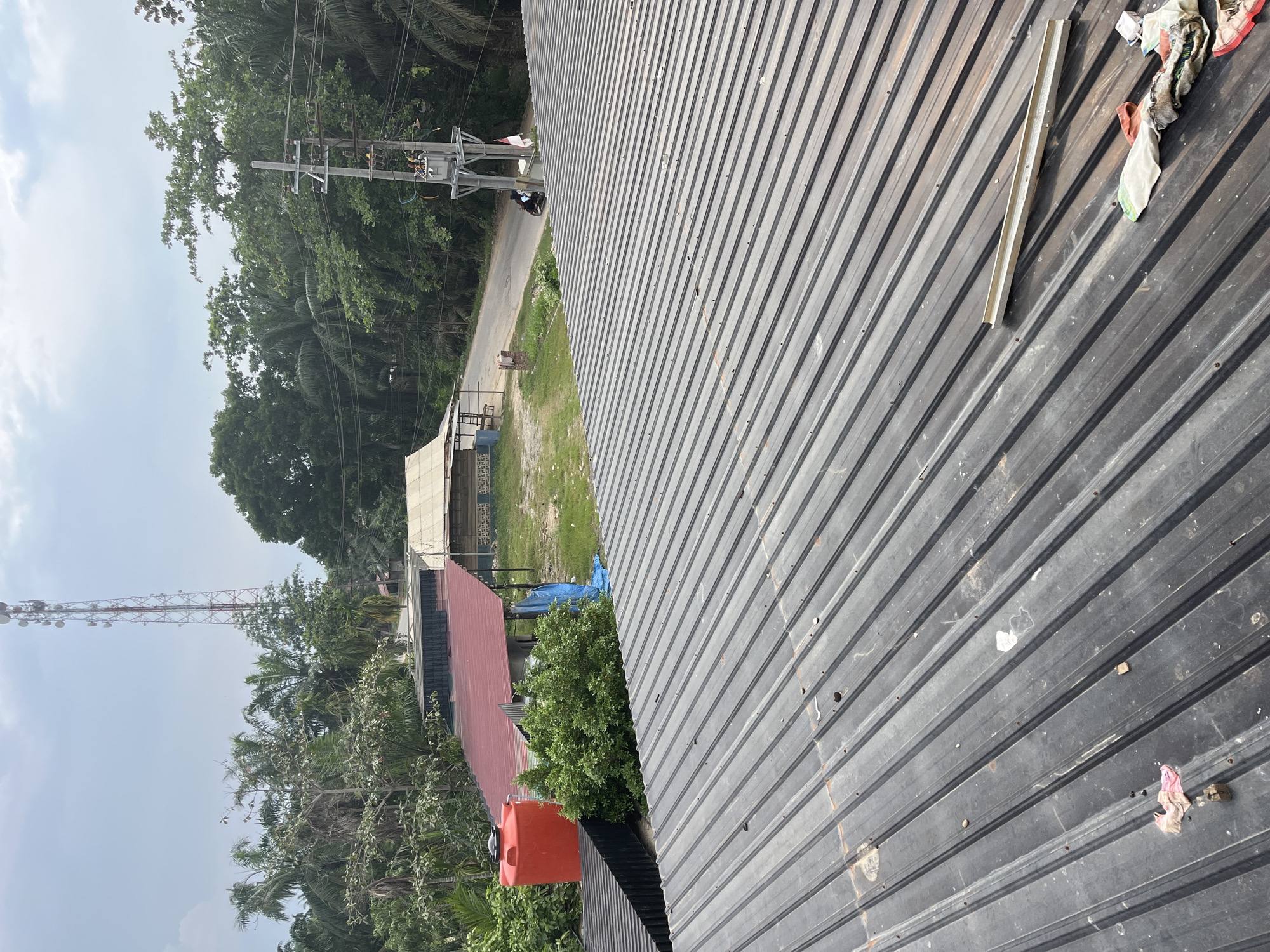 The image depicts a rooftop view of a semi-urban environment, possibly in Benuaraja, Indonesia. In the foreground, a dark gray corrugated metal roof dominates the scene, showing signs of wear and tear with scattered debris, including small pieces of wood and fabric.
Further into the background, a red water tank is visible, situated next to a building with a red-tiled roof. Beyond this, a road runs through the area, lined with green foliage and trees. A tall communication tower stands prominently in the distance, reaching towards a cloudy sky. A utility pole with electrical equipment and wires is also present, adjacent to the road. A person is seen riding a motorcycle on the road. The overall scene suggests a practical, functional setting, likely in a developing area.