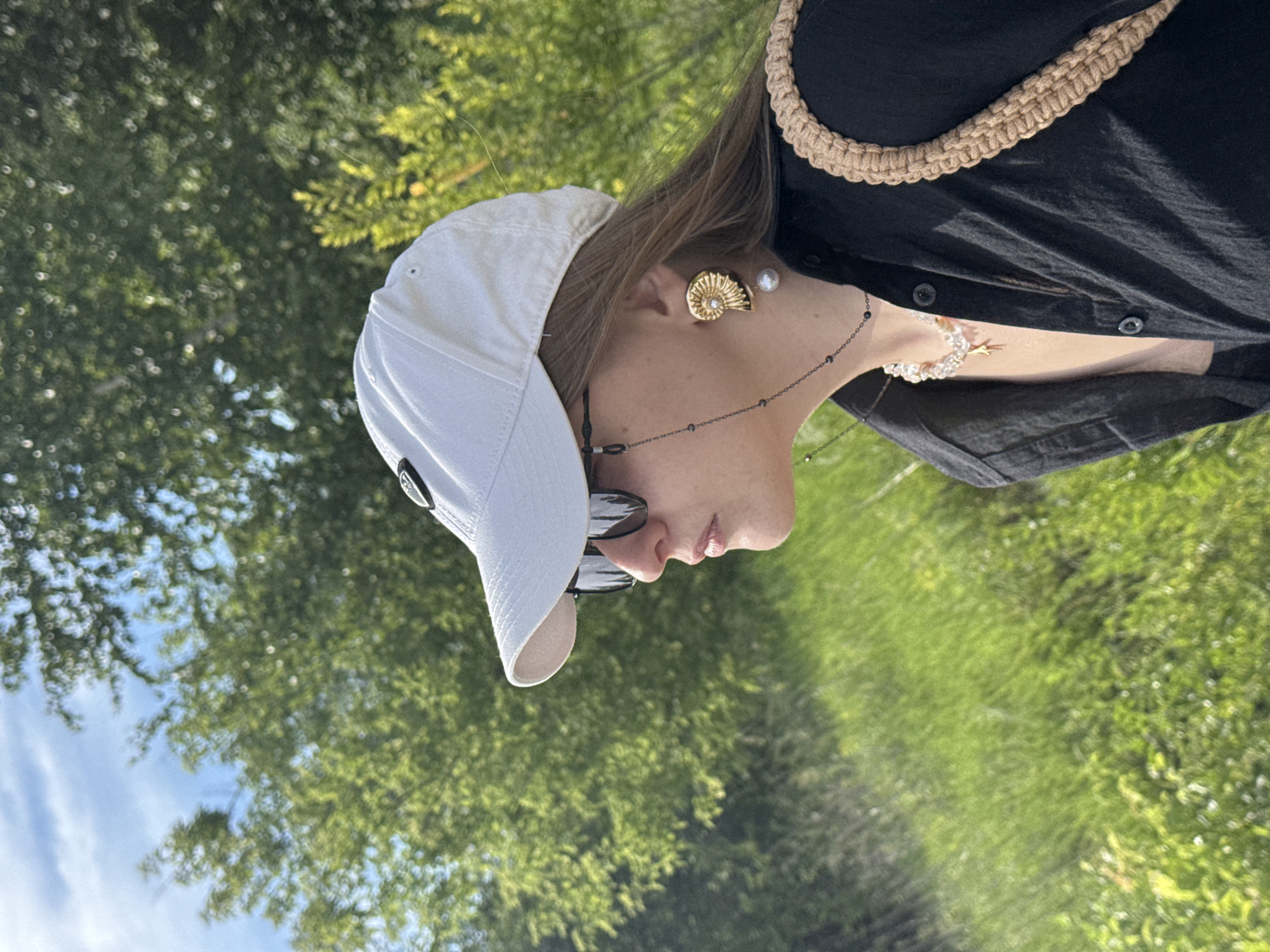 A medium close-up shot shows a person wearing a white baseball cap and sunglasses, facing left and looking off into the distance. The person's hair is brown and extends past their shoulders. They are wearing a black shirt with buttons down the front, a beaded necklace with a star pendant, and a unique gold earring shaped like a seashell with a pearl. A tan woven strap from a bag is visible over their right shoulder. The background is a blur of green foliage and grass, with a hint of blue sky and white clouds at the top left of the frame. The lighting suggests it is daytime, possibly late afternoon, with soft sunlight filtering through the trees. The scene appears to be outdoors, in a natural, grassy area with trees.