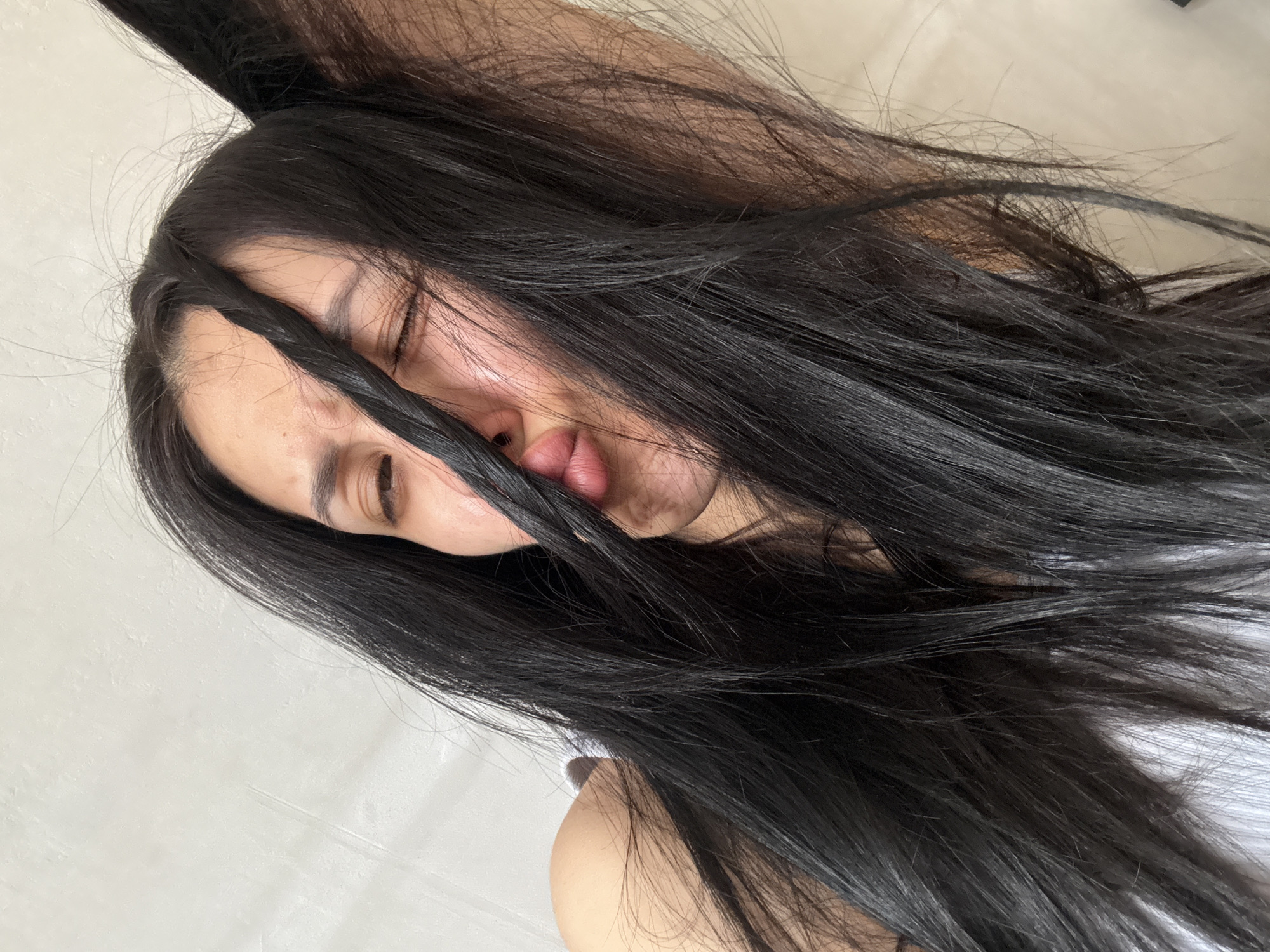 A woman with long, dark hair is lying down, possibly on a bed. Her eyes are closed. The background is a plain white or light-colored surface. The photograph is taken at an angle, with the camera positioned above the woman. There is no indication of Votkinsk, Russia or any specific location.