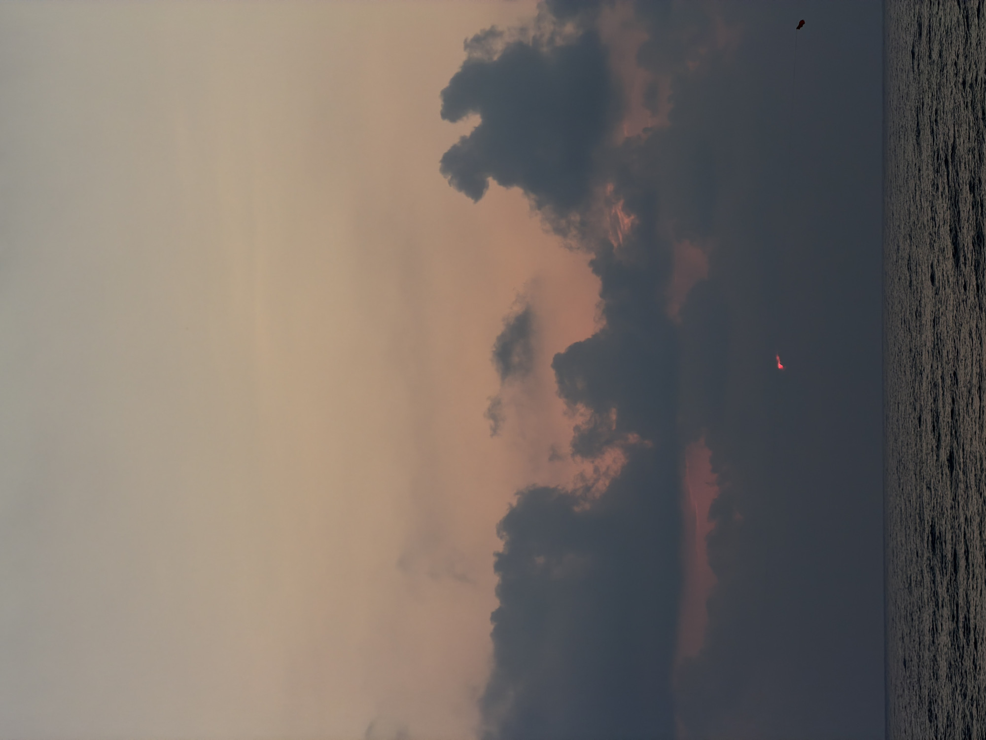 A sunset scene over the Mediterranean Sea, as viewed from Tel Aviv, Israel. The sky is filled with soft, gradient hues of orange, pink, and gray, transitioning from a lighter tone at the top to darker, more muted colors near the horizon. Dark, textured clouds dominate the middle and lower portions of the sky, with patches of warm light peeking through them, indicating the setting sun. A small, bright red point of light, likely the sun itself or a reflection, is visible low in the sky amidst the clouds. The sea in the foreground is dark and rippling, reflecting the dim light of the sky. In the far distance, on the right side of the frame, a small, dark object with a red tail is visible in the sky, possibly a kite or a drone.