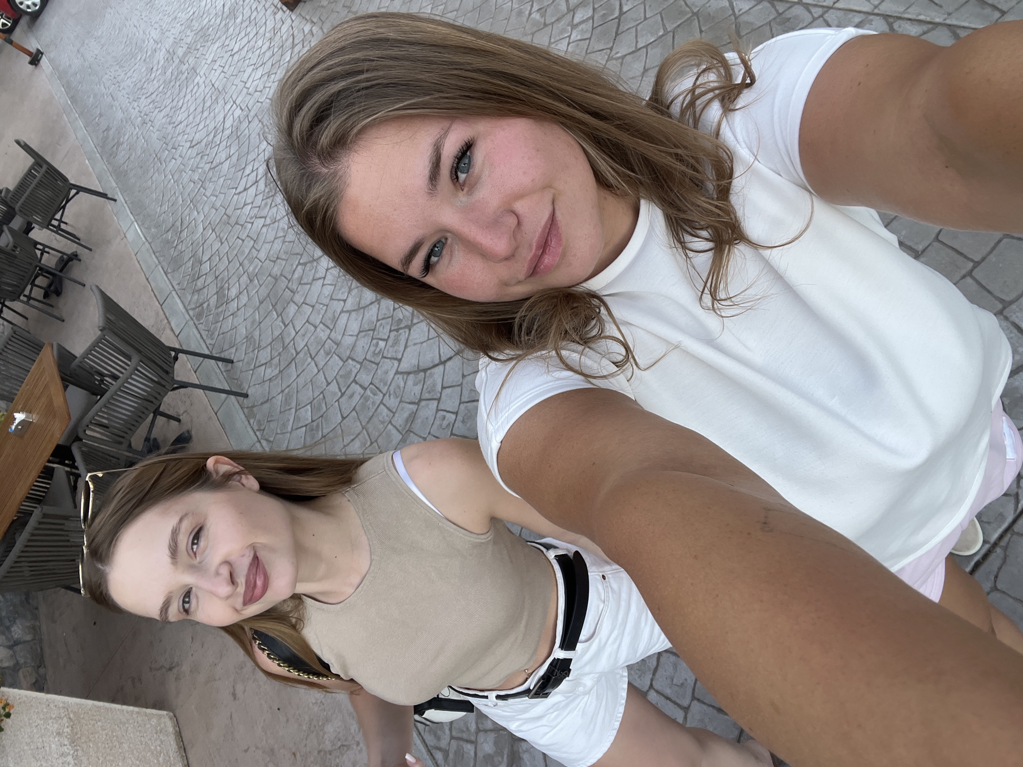 Two young women pose for a selfie on a stone-paved area, likely a patio or public space. The woman closer to the camera wears a white t-shirt, while the other wears a beige tank top and white shorts with a black belt. The background includes outdoor seating at a cafe or restaurant with tables and chairs. Based on the location query, this scene is likely in Kovačko Polje, Montenegro. There's no specific information to confirm the setting.