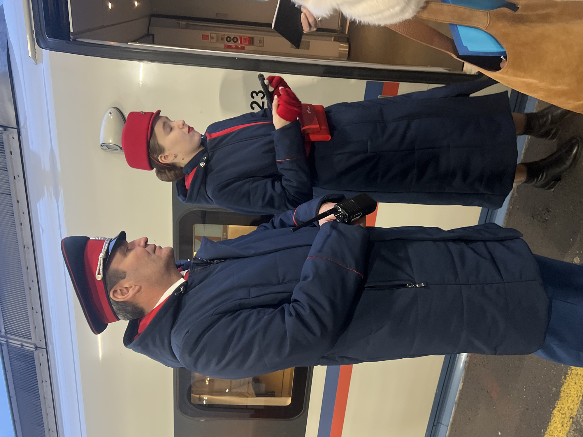 Two uniformed conductors stand next to an Amtrak train. The woman, in a red hat and gloves, is using a handheld device. The man wears a similar uniform with a peaked cap. The train is white with red and blue stripes. The location is not discernable from the image.