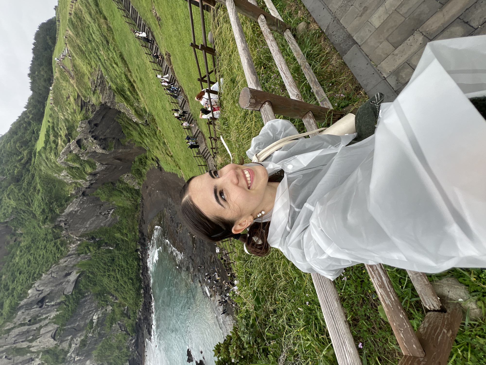 A woman is standing on a wooden structure overlooking a cliff and the ocean in Seogwipo-si, South Korea. She is wearing a white poncho and smiling at the camera. A boardwalk with people walking on it can be seen on the green, grassy hillside in the background. The ocean is turquoise with waves crashing against the rocky shore.