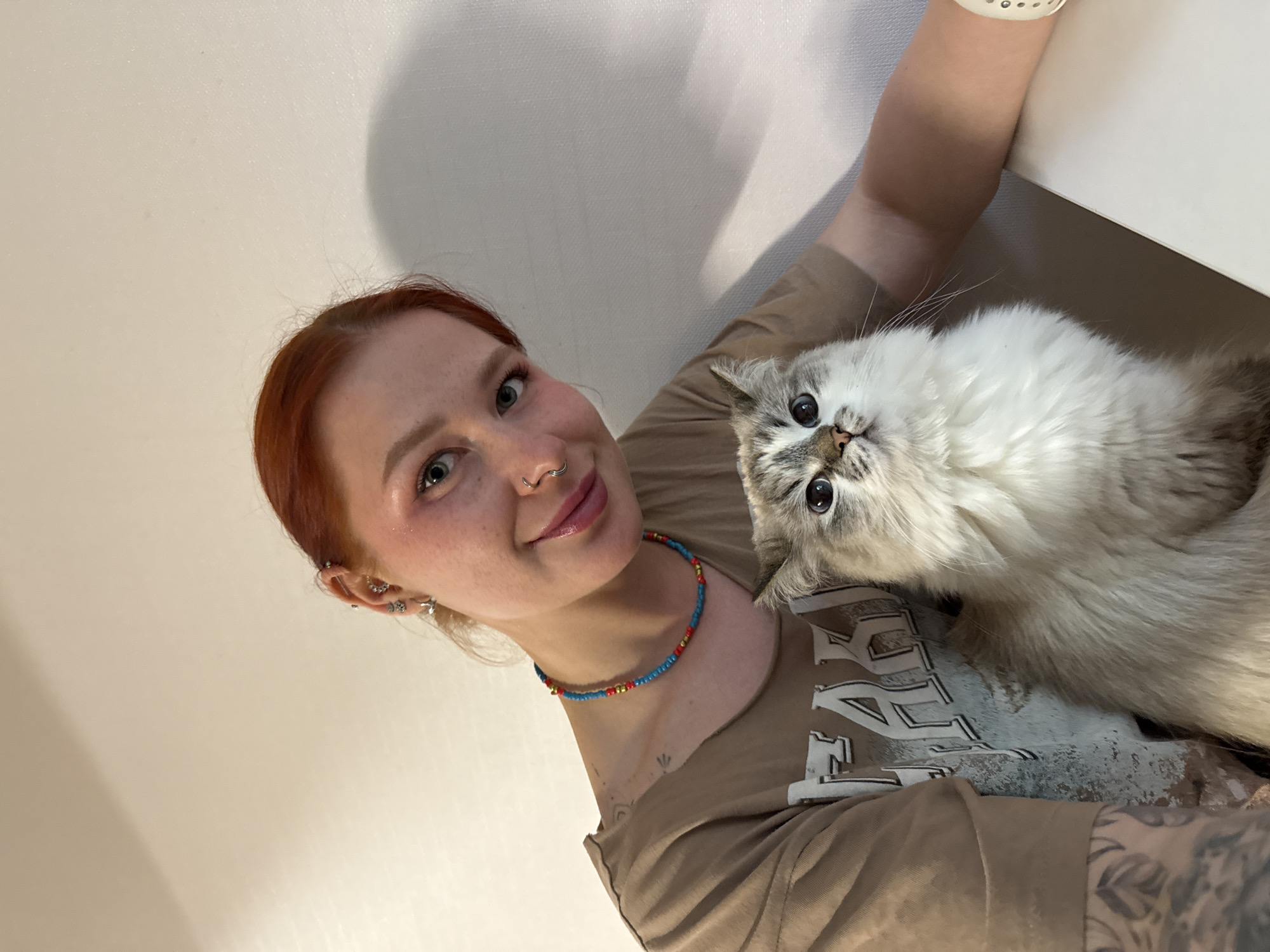 A selfie shows a fair-skinned woman with red hair and multiple ear piercings holding a fluffy, grey and white cat. The woman has a nose ring and is wearing a beaded blue, red, and yellow necklace. She is wearing a brown t-shirt with white lettering, partially obscured by the cat. The cat is looking directly at the camera with large, expressive eyes. The background consists of a plain, white wall and a partially visible white surface, possibly a table or counter. There's no evidence suggesting this image was taken in Rostov-on-Don, Russia.