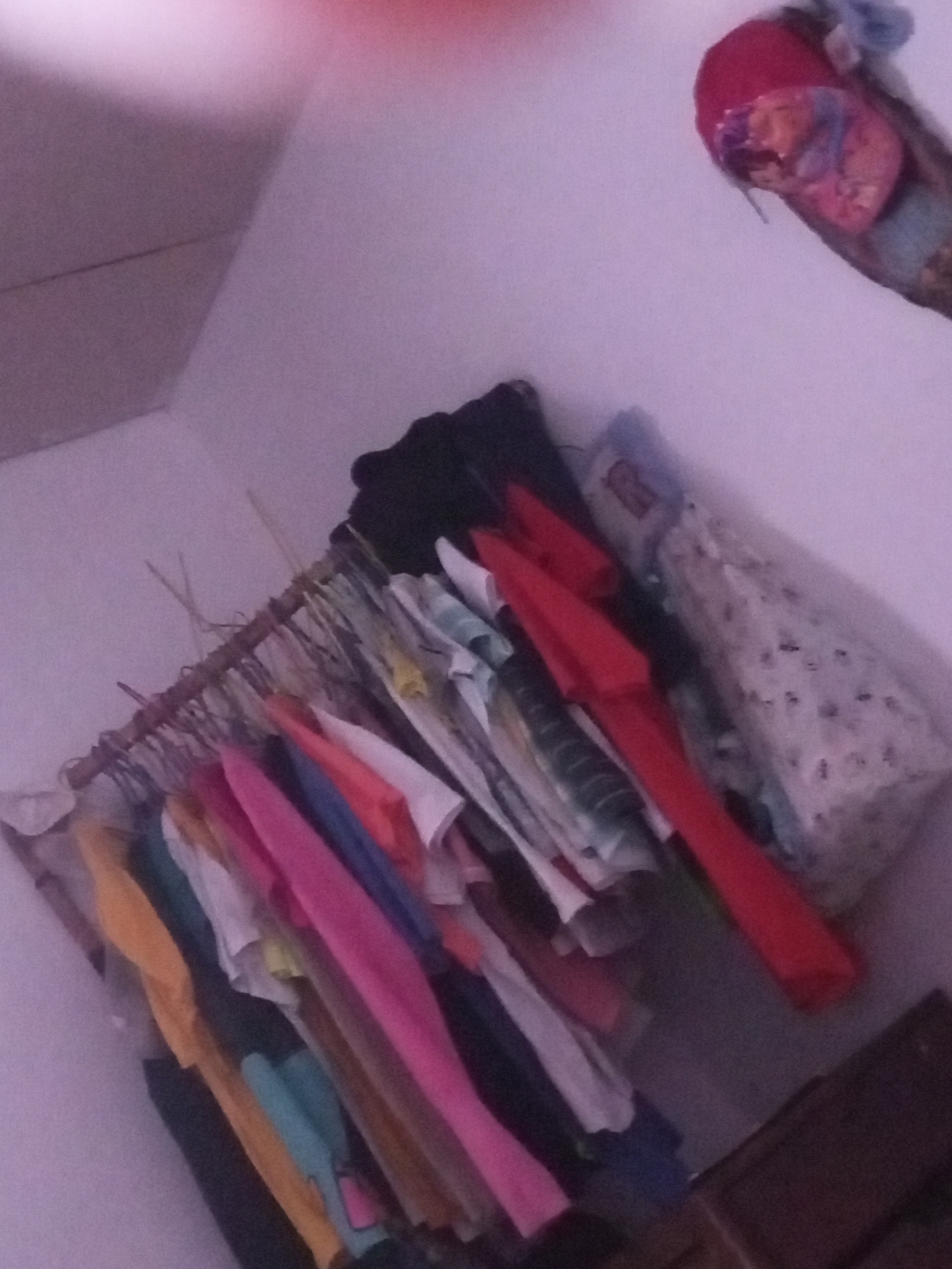 The image shows a closet or a similar storage space with clothes hanging on a makeshift clothes rack made of branches. The clothing items are of various colors including pink, blue, yellow, and white. There are also some folded items and a red object, possibly a hat or bag, at the top right of the frame. The background is a plain wall. The scene appears to be indoors, in what could be a residential setting in La Tagua, Venezuela. There are no people visible in the image.