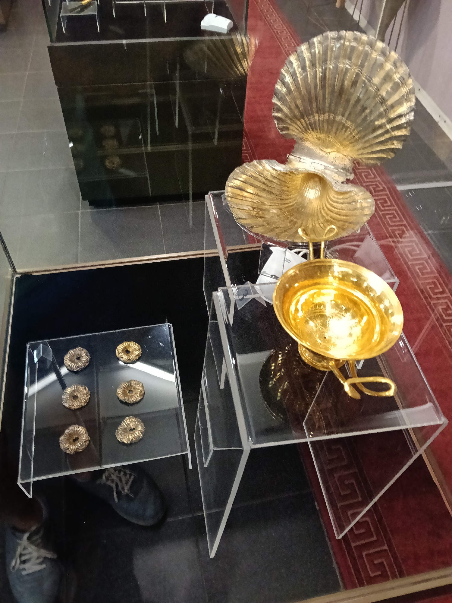 In a museum setting, likely in Kazanlak, Bulgaria, several golden objects are displayed. The primary subjects are ornate golden decorative items. A large, elaborate golden shell-shaped object is prominent, with a smaller, similarly shaped shell beneath it. A golden bowl on a stand, also with decorative elements, is positioned on a clear acrylic display. To the left, on another clear acrylic stand, are four smaller circular golden objects, each with intricate, textured designs. These objects are displayed in glass cases. The overall scene suggests an exhibition of valuable artifacts. A red carpet with a Greek key pattern is visible in the background, adding to the formal display. A pair of blue sneakers are visible, reflecting in the glass, indicating the presence of a person or people observing the exhibit. The lighting is indoor, suggesting daytime or artificial lighting suitable for a museum. There is no visible text in the image.