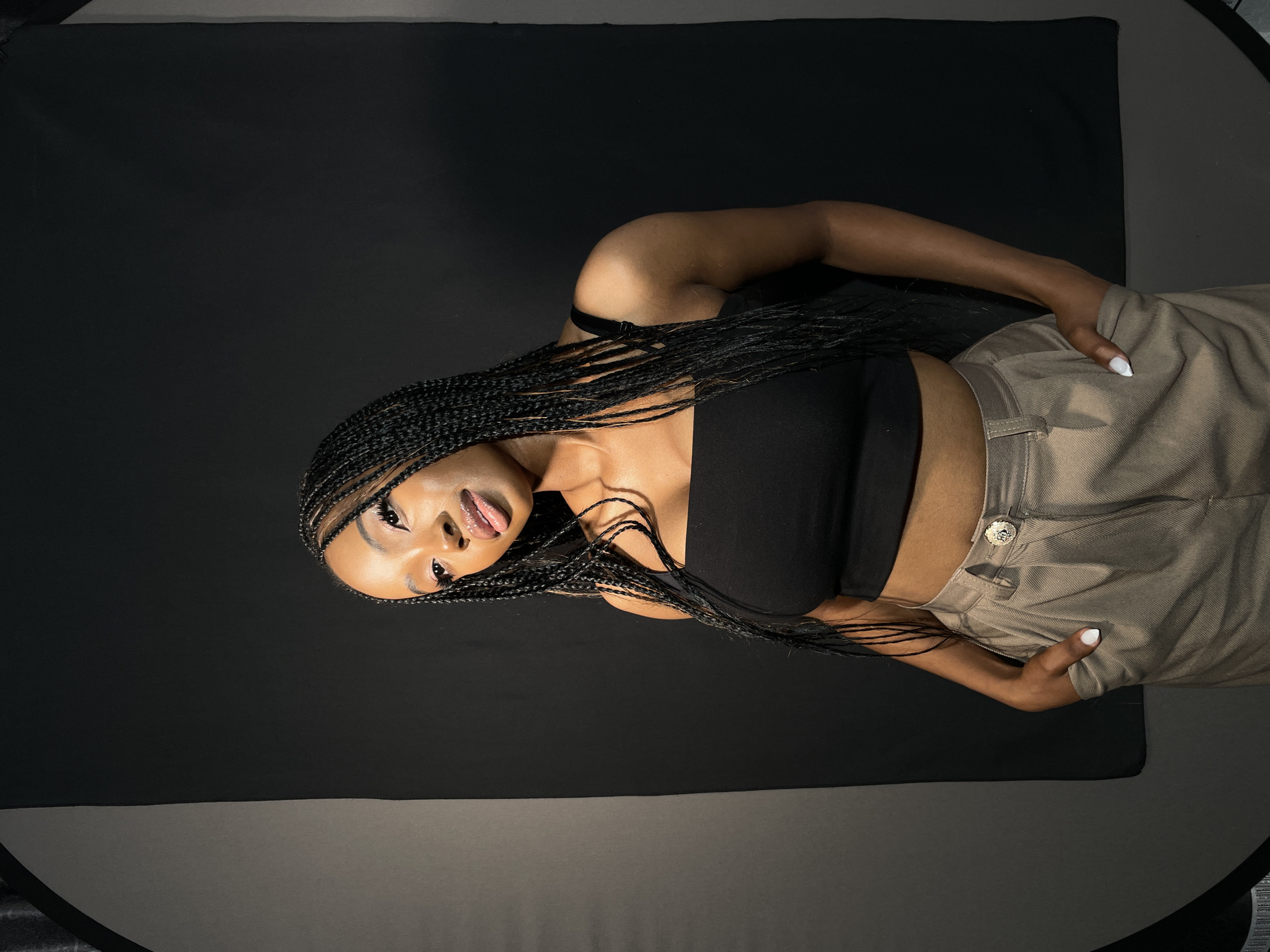 A medium shot captures a young Black woman posing in what appears to be a photography studio. She is positioned on a dark background, with her head tilted to the right and her body angled diagonally across the frame. Her long, black braided hair cascades over her shoulders and chest. She is wearing a black crop top that exposes her midriff and light brown or khaki-colored pleated pants with a distinct silver-toned button at the waist. Her left hand is subtly placed near her hip, while her right arm is bent.
The woman's face is prominent, featuring winged eyeliner, defined eyebrows, and glossy lips, with her tongue slightly out, conveying a playful or confident expression. Her fingernails are painted white. The lighting highlights her features and the texture of her braids. The background is predominantly dark black, transitioning to a dark gray surface at the bottom of the frame, suggesting a plain studio backdrop. There is no visible text or specific location cues beyond the studio setting.