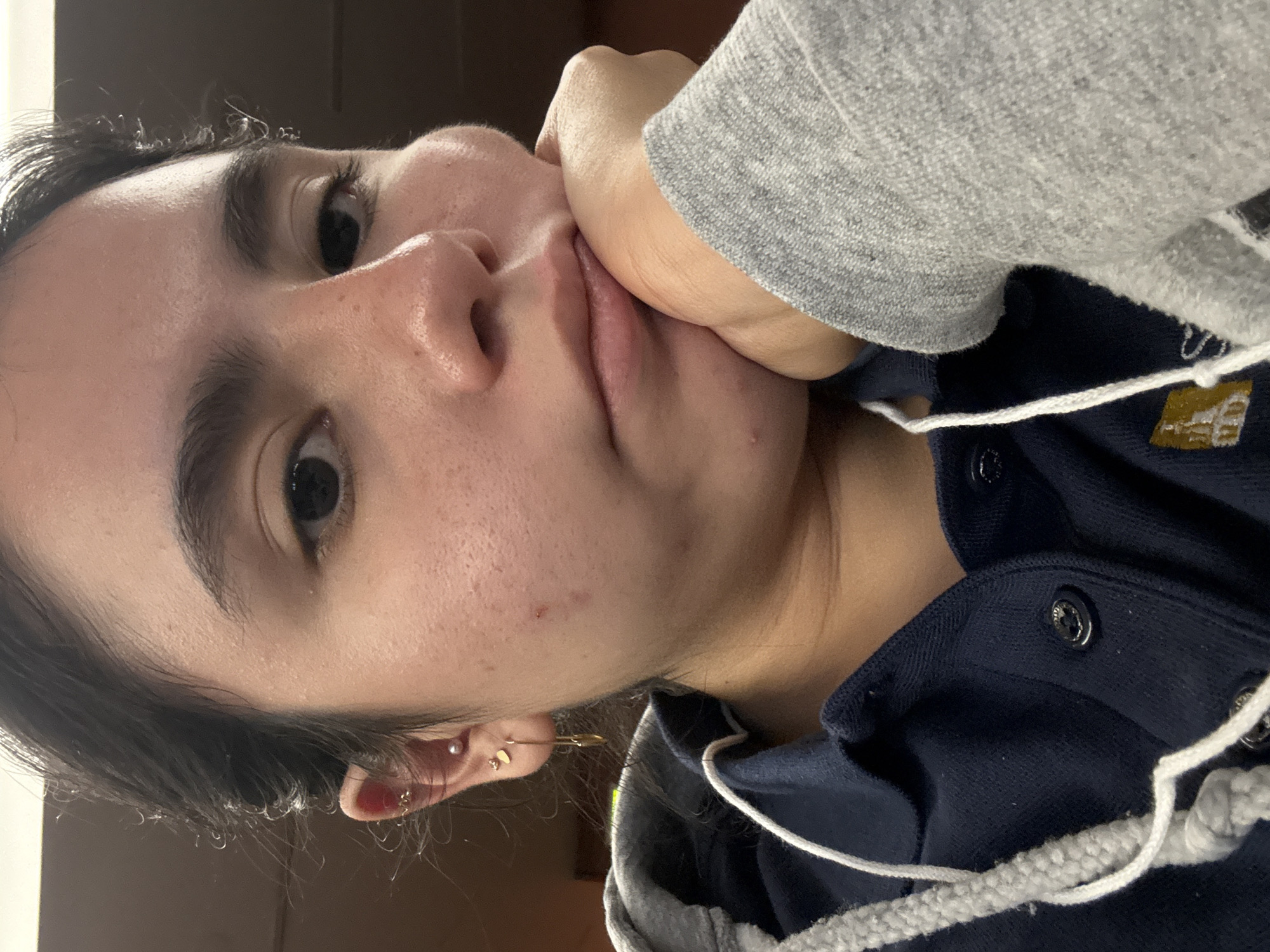 A close-up, angled photograph shows a person with dark hair, light skin, and multiple ear piercings. The individual's face is partially obscured by their hand, which rests against their cheek. The person is wearing a blue jacket with a gray hood and white drawstrings. The background is indistinct, appearing to be an indoor setting with neutral tones. There is no specific information within the image that would indicate the location is in San Antonio, United States of America.