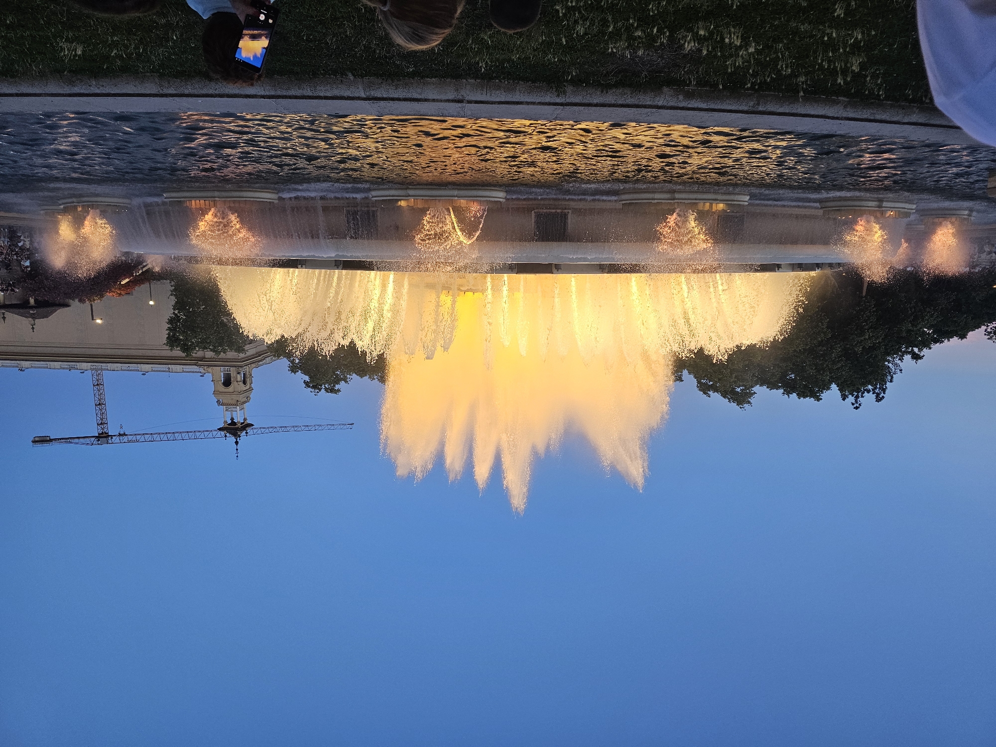 The image depicts a wide shot of the Magic Fountain of Montjuïc in Barcelona, Spain, at dusk. The fountain is illuminated with bright yellow and orange lights, creating a spectacular display of water jets. In the background, a tower with a spire and a construction crane are visible, as well as trees and buildings. Several people are present, some are seated on the grass in the foreground, and one person is holding a phone to take a picture of the fountain. The sky is a deep blue, indicating the time of day.