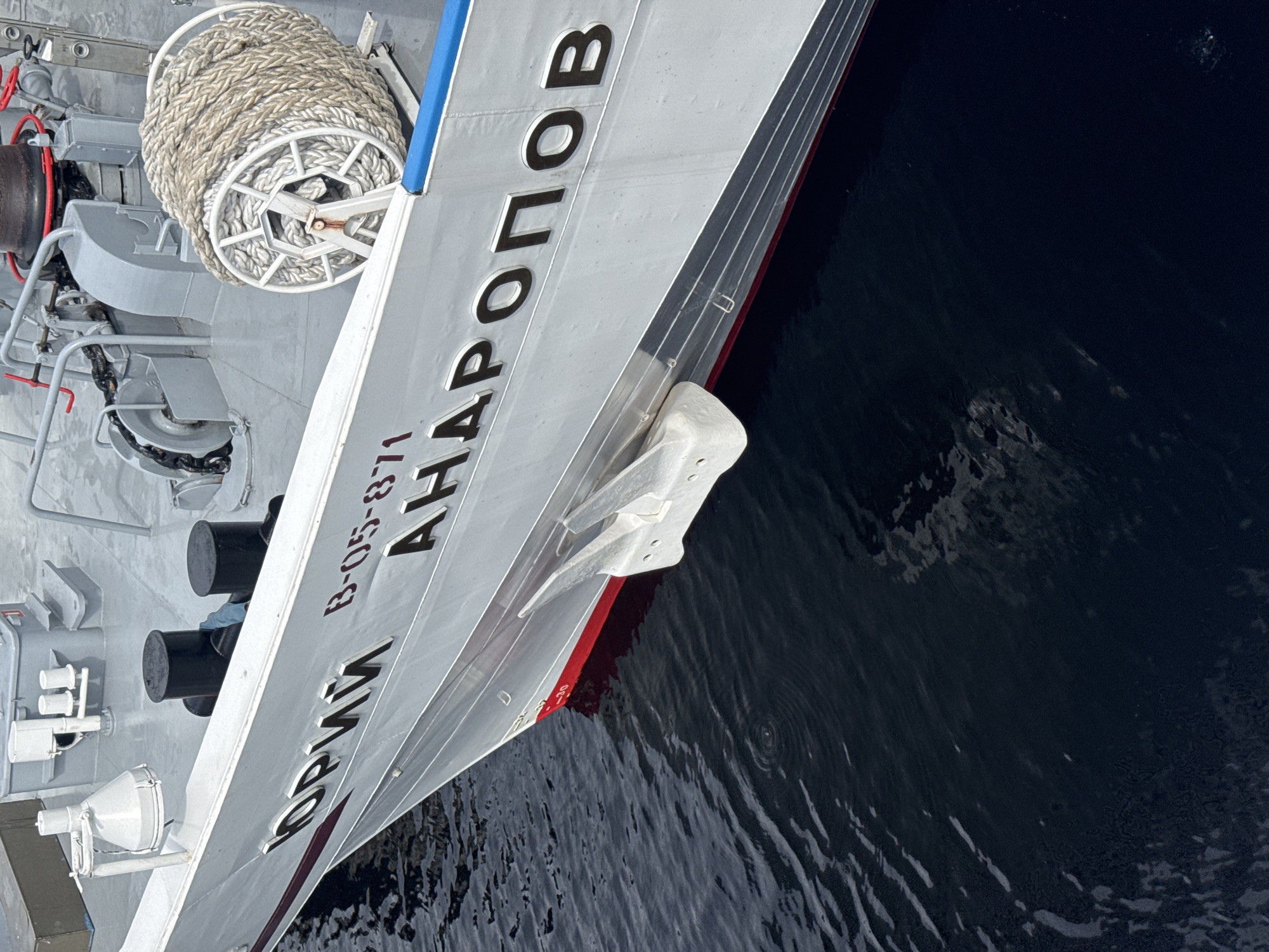A close-up, angled shot shows the side of a white ship docked in dark water. The ship has a red stripe along its waterline. Bold, dark lettering on the ship's hull reads "ЮРИЙ АНАПРОВ" and "B-05-871". To the left, a large spool of thick rope is visible, along with various deck equipment, including black bollards and white lights. The water is dark and choppy, with some reflections of the sky. The scene appears to be set during the daytime, with overcast or neutral lighting conditions. The image is taken from a slightly elevated perspective, looking down at the ship and the water. The setting is likely a harbor or port, given the presence of a docked vessel and deck equipment. There are no people visible in the image.