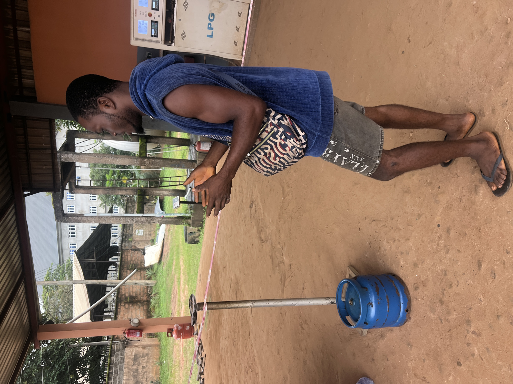 In Nkpor, Nigeria, a man stands in a covered outdoor area with a dirt floor. He is wearing a blue sleeveless hoodie, grey shorts, and flip-flops. A patterned crossbody bag is slung over his shoulder. In front of him, a metal pole is secured to the ground by a blue propane tank. A pink tape stretches horizontally across the scene, held by the man's hands. To the left, a red fire extinguisher is mounted on a pole. In the background, a white LPG gas pump with digital displays stands near a wall. Further back, there is a brick wall, lush green grass, and a multi-story building with many windows.