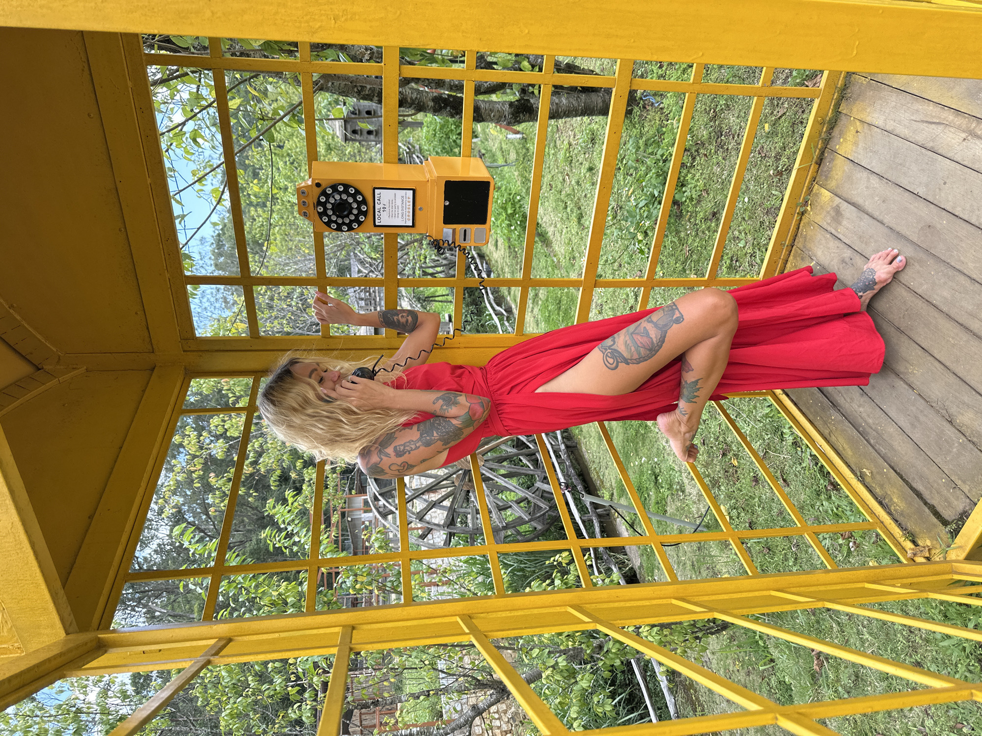 In a bright yellow, open-air structure resembling a phone booth, a woman with blonde, wavy hair stands on a wooden floor. She is wearing a vibrant red dress with a high slit on the left side, revealing her tattooed legs. The woman is holding a black telephone receiver to her ear and appears to be speaking on it. Her left hand rests on the yellow frame of the booth, and her right arm is extended towards the phone.
The phone itself is a vintage-style yellow model, mounted on the wall of the booth. It has a rotary dial and a slot for coins, with text that reads "LOCAL CALL 10 / 7" and "LONG DISTANCE".
The background of the scene is lush and green, suggesting an outdoor setting. Trees with abundant foliage, grass, and a glimpse of a rustic wooden water wheel are visible through the open structure of the booth. The environment is natural and somewhat untamed, with a few man-made elements like a birdhouse structure in the background. The overall impression is a quirky and colorful scene set within a natural landscape. While the provided location is Cam Ly - Đà Lạt, Vietnam, there are no specific visual cues in the image that directly confirm this location.