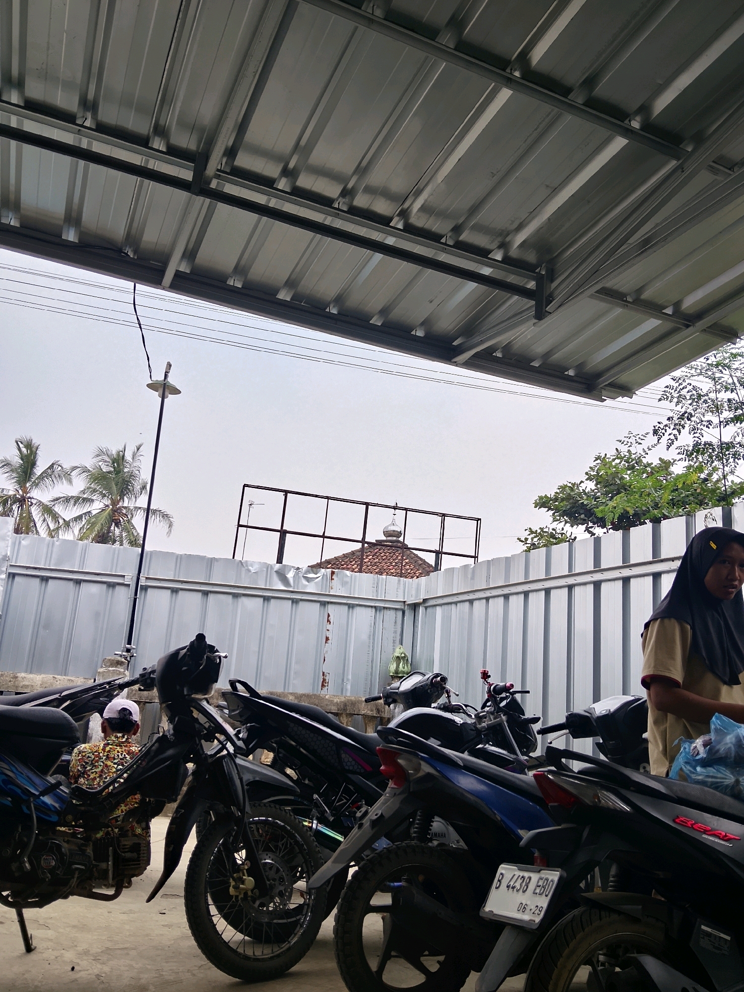 The image depicts an outdoor scene beneath a corrugated metal roof structure. Several motorcycles are parked in a row, some in the foreground and others further back. A woman wearing a headscarf and a light-colored shirt is standing to the right, looking towards the viewer. To the left, a person wearing a camouflage-patterned shirt and a white cap is seated on a motorcycle. In the background, a light-colored metal fence runs horizontally, with palm trees visible beyond it. A tall pole with a lamp fixture is also present, and further back, a building with a domed roof and a metal frame structure are visible. The sky is overcast.