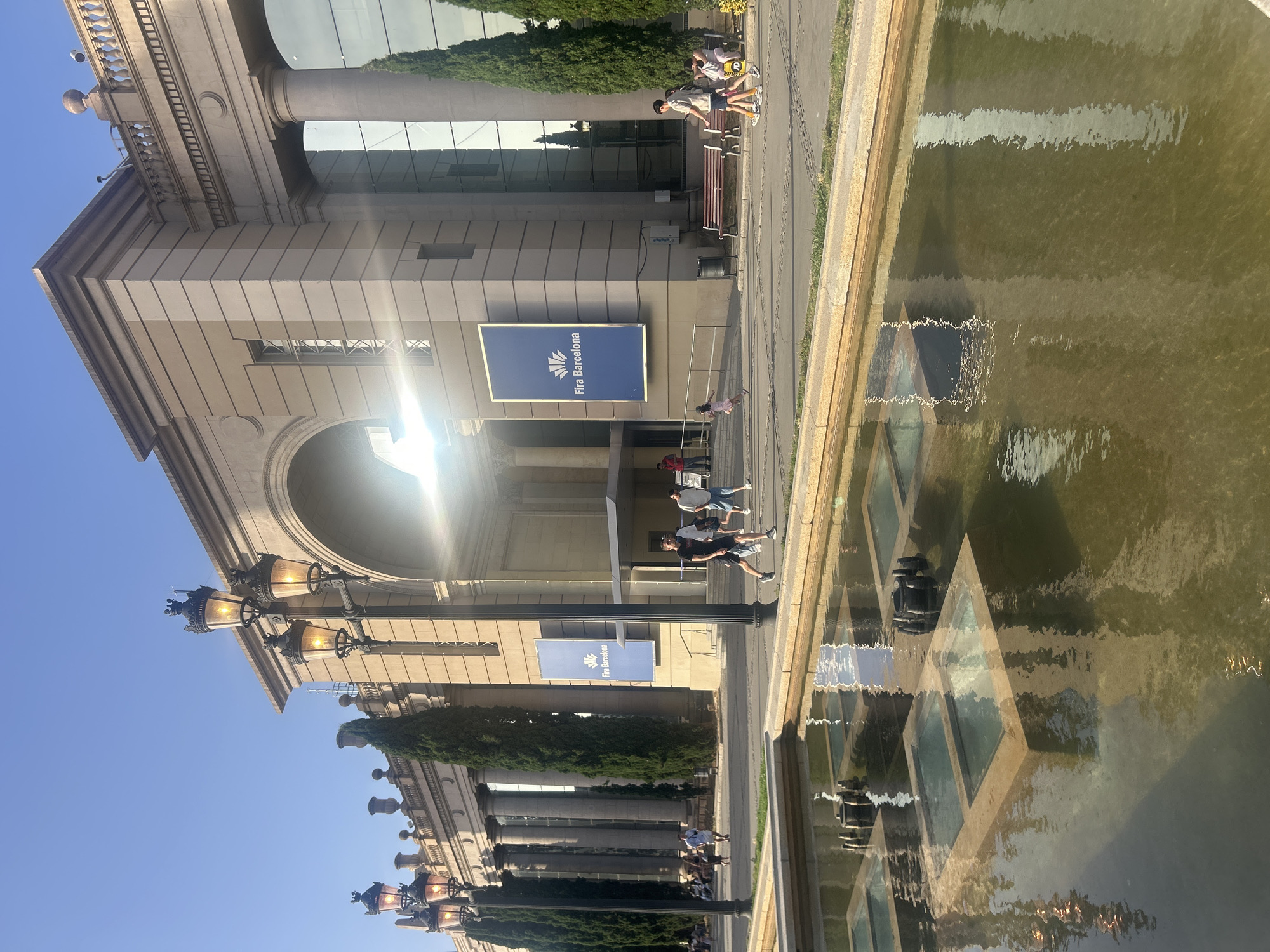 The image captures an outdoor scene in front of a large, neoclassical building in Barcelona, Spain, identified by a banner reading "Fira Barcelona". The building features a grand arched entrance and is flanked by rows of tall columns, with large glass windows reflecting the sky and surrounding trees. In the foreground, a decorative fountain with multiple square water features is visible, its murky water reflecting the building and sky. Several people are walking and standing in front of the building, some in groups and others individually. A streetlamp with multiple globes stands prominently on the left side of the image. The overall environment suggests a public space on a sunny day, likely a convention center or exhibition hall.