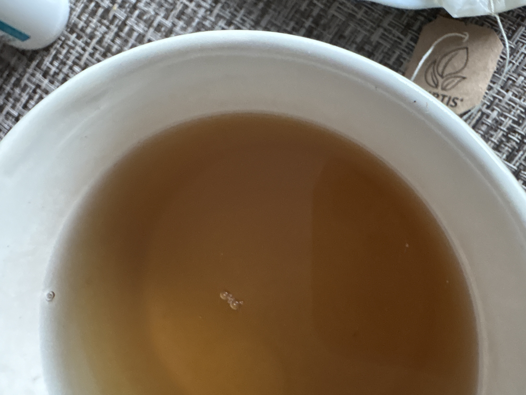 A close-up, top-down shot depicts a white bowl filled with a light brown liquid, likely tea. A tea bag with the word "RTIS" visible on its tag rests on the edge of the bowl. Small bubbles are present on the surface of the liquid. The bowl sits on a textured, woven surface. The location of Прикамский, Russia, is not relevant to the description of the objects and details visible in the image.