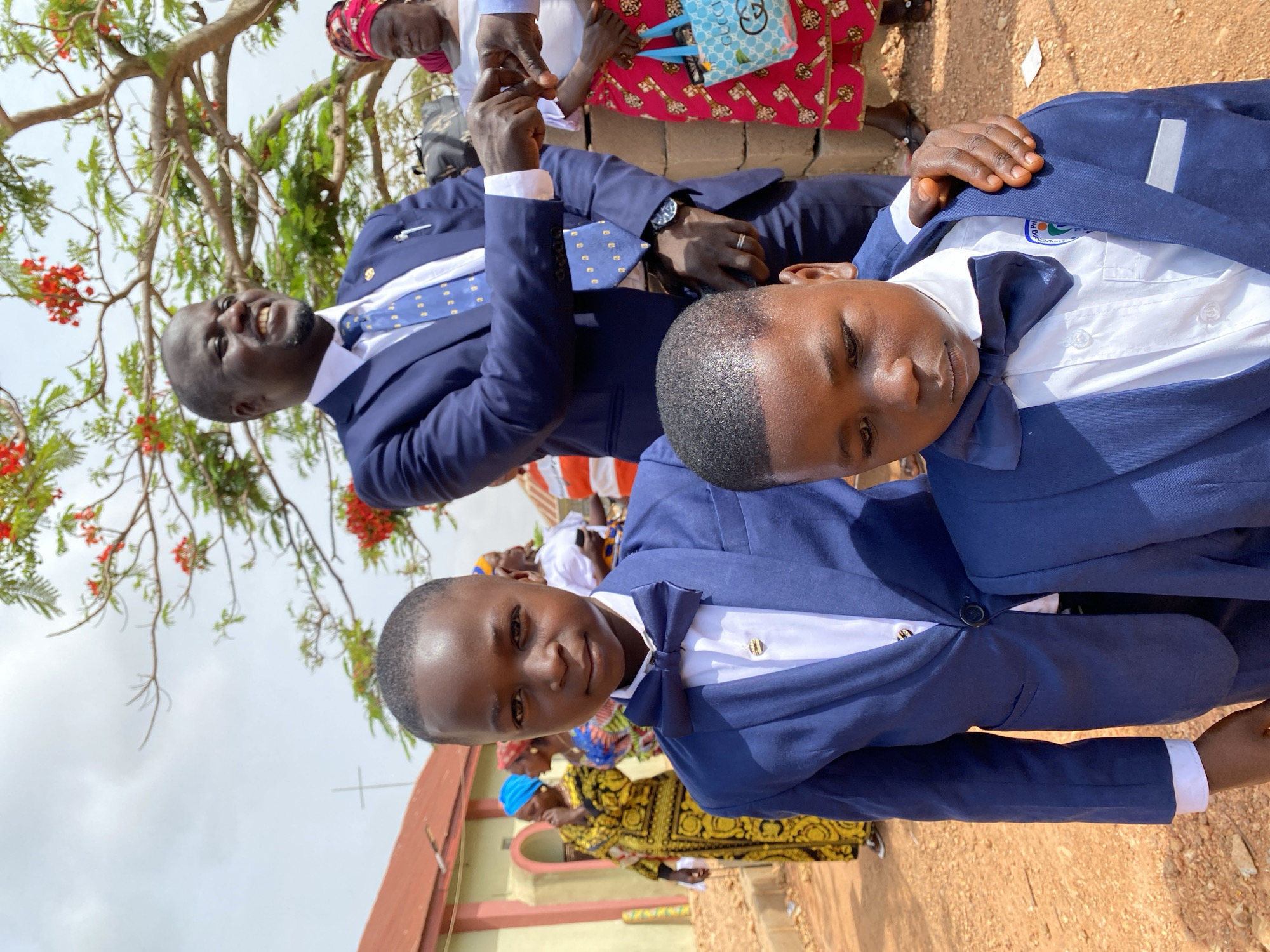 The image captures a scene in Kuru Dungus, Nigeria, featuring two young boys and an adult man. All three are formally dressed in navy blue suits with white shirts and ties or bow ties. The man stands behind and partially above the boys, his hand touching one of the boy's shoulder. The boys are standing close together, with the boy in the foreground looking directly at the camera and smiling slightly, while the other has his eyes cast downward. The man is smiling, his left arm raised slightly as if in a greeting gesture.
The background is filled with other individuals, some wearing colorful, patterned clothing and head coverings, and a building with a cross on its roof, suggesting a church. A tree with red flowers is visible in the upper left corner of the frame. The ground is a reddish-brown color, possibly dirt.