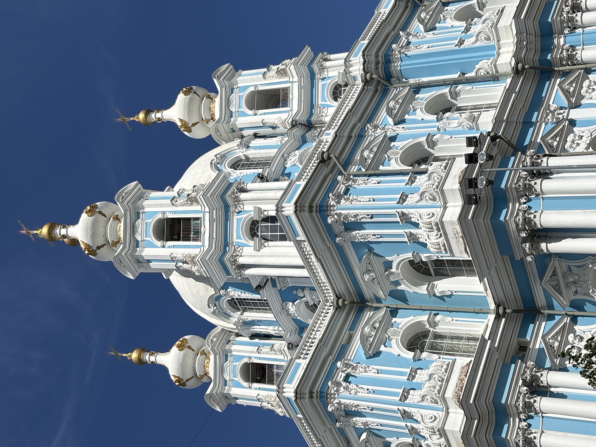 The image shows the Smolny Cathedral, a Russian Orthodox cathedral located in Saint Petersburg, Russia. The cathedral is a prominent landmark known for its distinctive blue and white Baroque architecture. The image focuses on the upper portion of the cathedral, highlighting its ornate domes, gilded crosses, and intricate decorative details. The bright blue sky serves as a backdrop, emphasizing the vibrant colors of the building. There are no people visible in the image. The scene is a close-up, low-angle shot of the cathedral's facade.