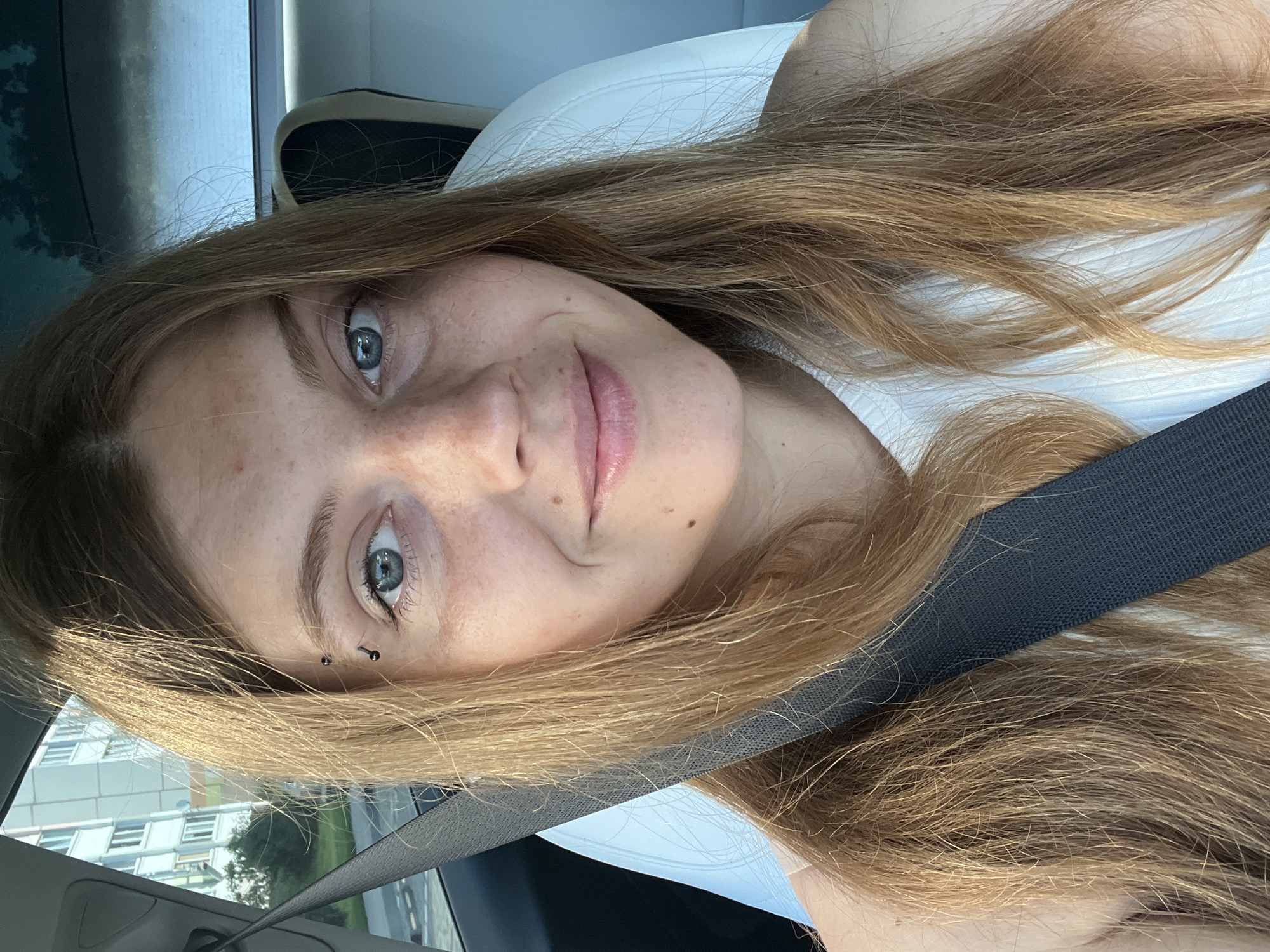 A young woman with long, light brown hair and blue eyes is sitting in the passenger seat of a car. She is wearing a white top and has a seatbelt fastened across her chest. She has freckles on her face and a small black eyebrow piercing. The background shows a blurry view of a building and some trees, suggesting an outdoor urban setting. The scene appears to be in the city of Senitsky Sel'skiy Soviet, Belarus.