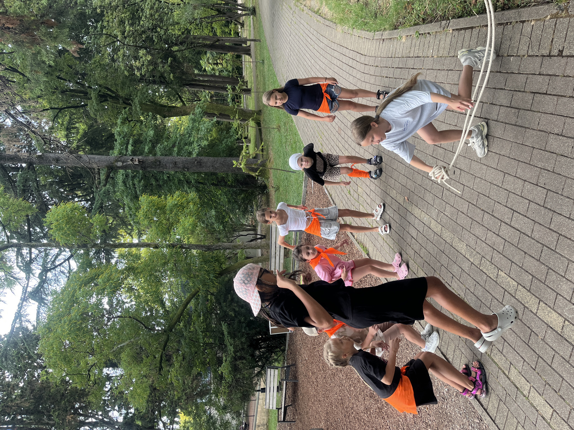 A group of seven children, appearing to be of elementary school age, are engaged in an outdoor activity in a park setting. The children are dressed in casual, warm-weather clothing, with some wearing bright orange sashes or accessories. The ground beneath them is paved with a light-colored brick pattern, and the park is surrounded by lush green trees and foliage. In the background, a wooden bench and a mulched area are visible, suggesting a recreational or educational environment. The children's poses and expressions indicate they are actively participating in a game or exercise. One child in the foreground is mid-jump, wearing a black t-shirt, black shorts with an orange sash, and a pink patterned cap. Another child on the right is crouching and holding a rope, appearing to be initiating or participating in a rope-related activity. The overall scene suggests a summer day spent outdoors, possibly during a camp or organized event.
