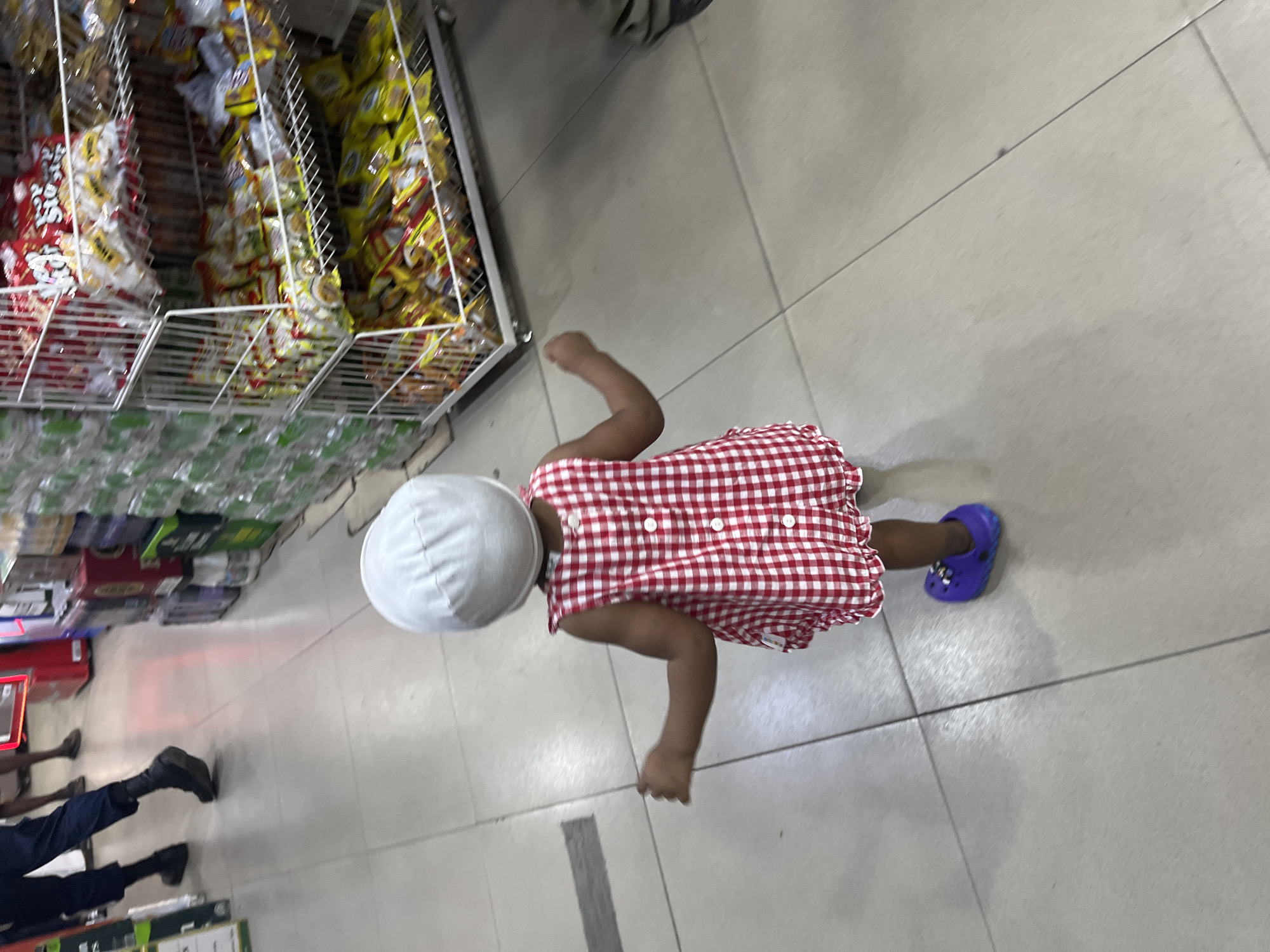 The image depicts a child in Ado, Nigeria, lying on the tiled floor of a grocery store. The child wears a white hat, a red and white checkered dress, and purple Crocs. The child is near a shelf stocked with snacks and beverages. Two individuals wearing black shoes and dark pants can be seen in the lower-left corner. The focus is on the child.