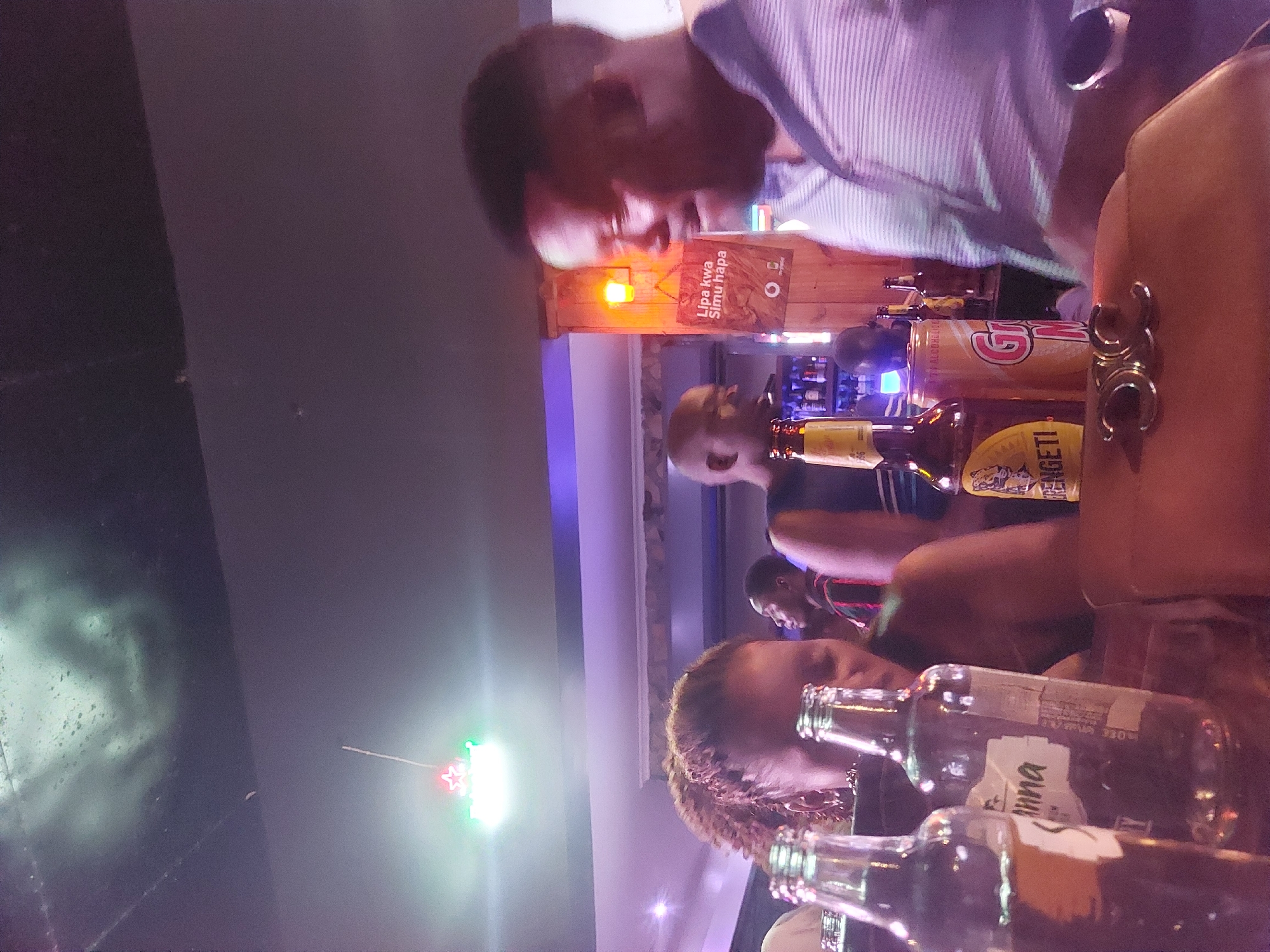 A dimly lit bar in Dar es Salaam, Tanzania, shows four patrons gathered around a table covered with empty beer bottles and a can of soda. A man wearing a striped shirt stands on one side of the table. The bar has a wooden structure with a sign that reads "Lipa kwa Simu hapa" (Pay by phone here). A string of Christmas lights hangs from the ceiling, and liquor bottles sit on shelves behind the bar.