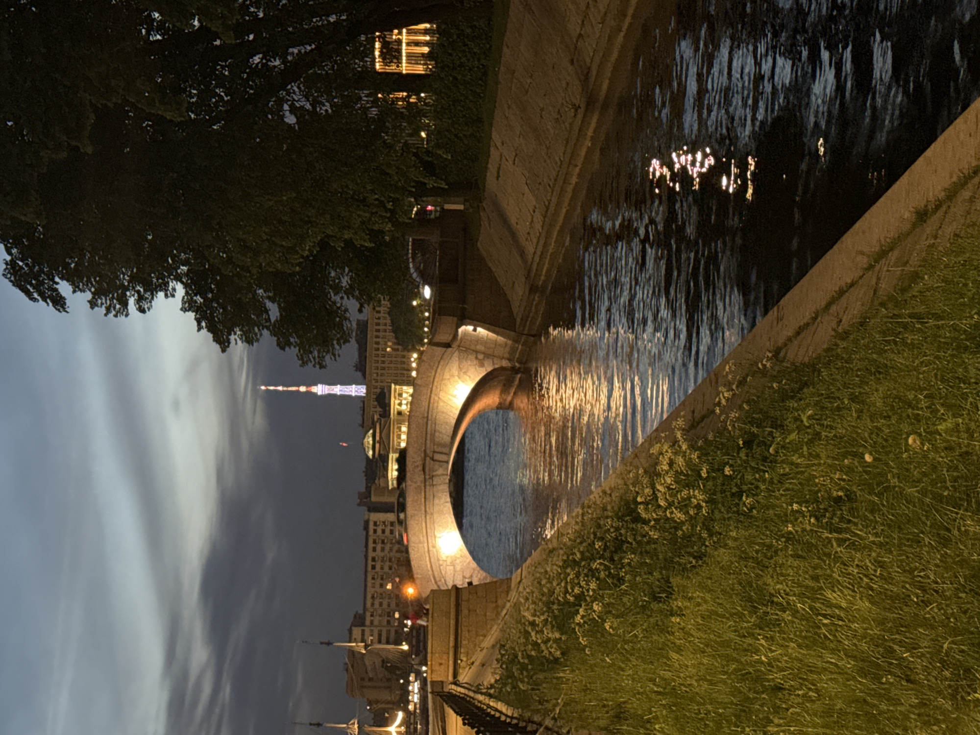 This image depicts an evening scene along the Moika River in Saint Petersburg, Russia, looking generally east-northeast towards the Saint Petersburg TV Tower.
In the foreground, a verdant grassy slope with scattered small white flowers descends towards a concrete embankment wall bordering a dark, rippled canal. A black metal fence runs along the top of the embankment on the left. The water reflects warm yellow light from nearby sources and scattered white and pink light from the distant city.
In the midground, a single-arch, light-colored stone bridge spans the canal. The bridge's arch is illuminated from below with warm yellow light, creating strong reflections in the water. A dark vehicle is partially visible on the bridge deck, moving from left to right. On the left embankment, just before the bridge, a person wearing a red top stands near the water.
The background features a cityscape under a dark, cloudy twilight sky. Buildings with illuminated windows are visible behind the bridge. To the far left, the masts and rigging of a large sailing ship are discernible. Dominating the distant skyline is the slender, illuminated Saint Petersburg TV Tower, displaying segmented bands of white, blue, and reddish-pink light. On the right side of the frame, large, dark trees obscure parts of the sky and buildings. Further down the canal on the right, another concrete embankment wall and a dark hedge are visible, with bright, vertically-oriented golden light reflections in the water.