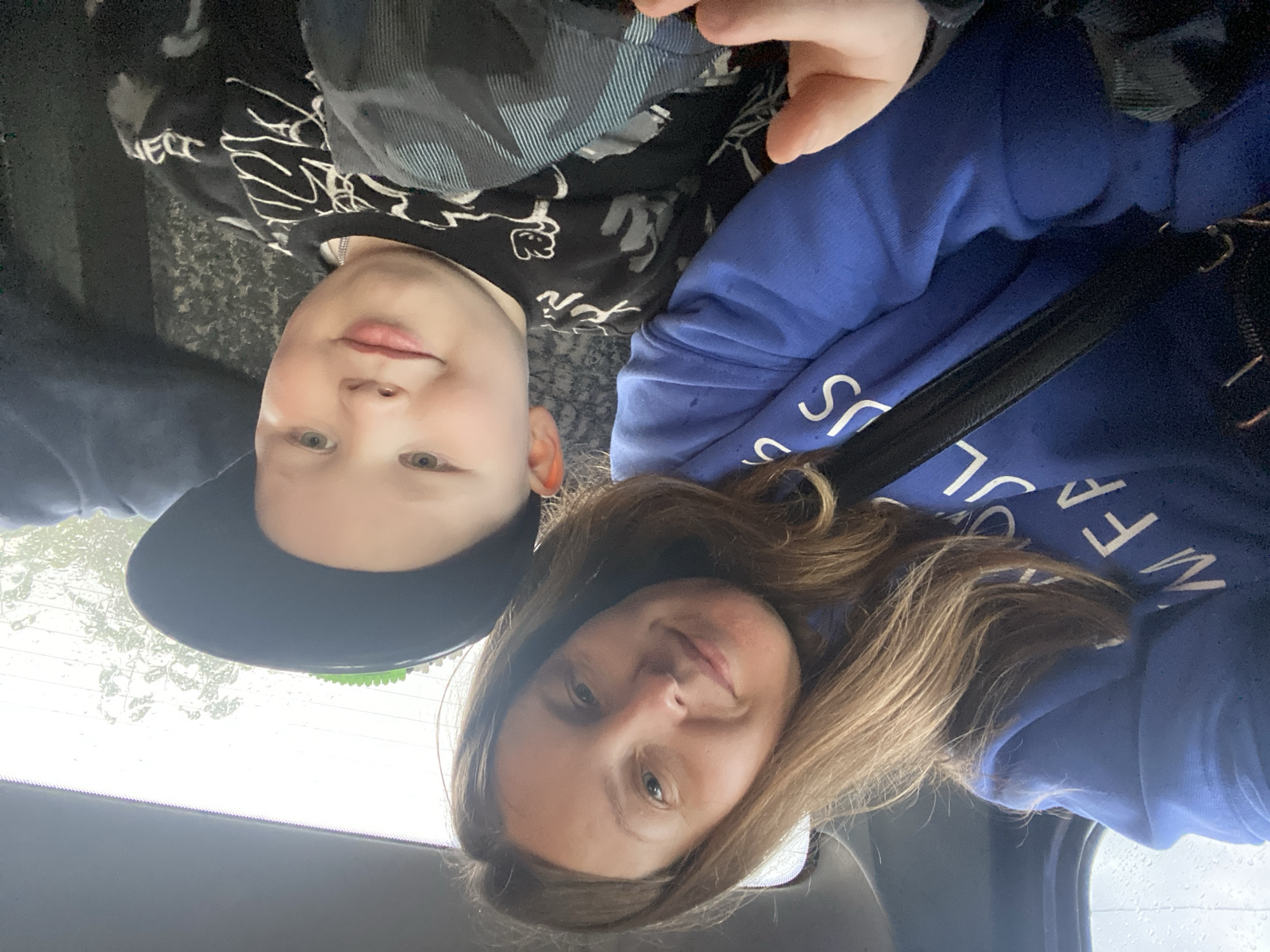 This image, originally rotated 90 degrees counter-clockwise, presents a selfie of an adult woman and a young child, likely a boy, inside a vehicle. The woman, positioned on the right with long, light brown hair, wears a blue hoodie displaying partial white text, including "US" and other fragments. She has a gentle, content smile. On the left, the child, wearing a black baseball cap with a visible green brim, a black patterned long-sleeved shirt with white designs, and a silver chain, also smiles subtly towards the camera. Both subjects convey a calm and pleasant mood. Behind them, a window reveals blurred outdoor surroundings with what appears to be raindrops or condensation, indicating an overcast or rainy daytime. The interior suggests they are in a car or similar vehicle. The provided location context is Chaikovsky, Russia.