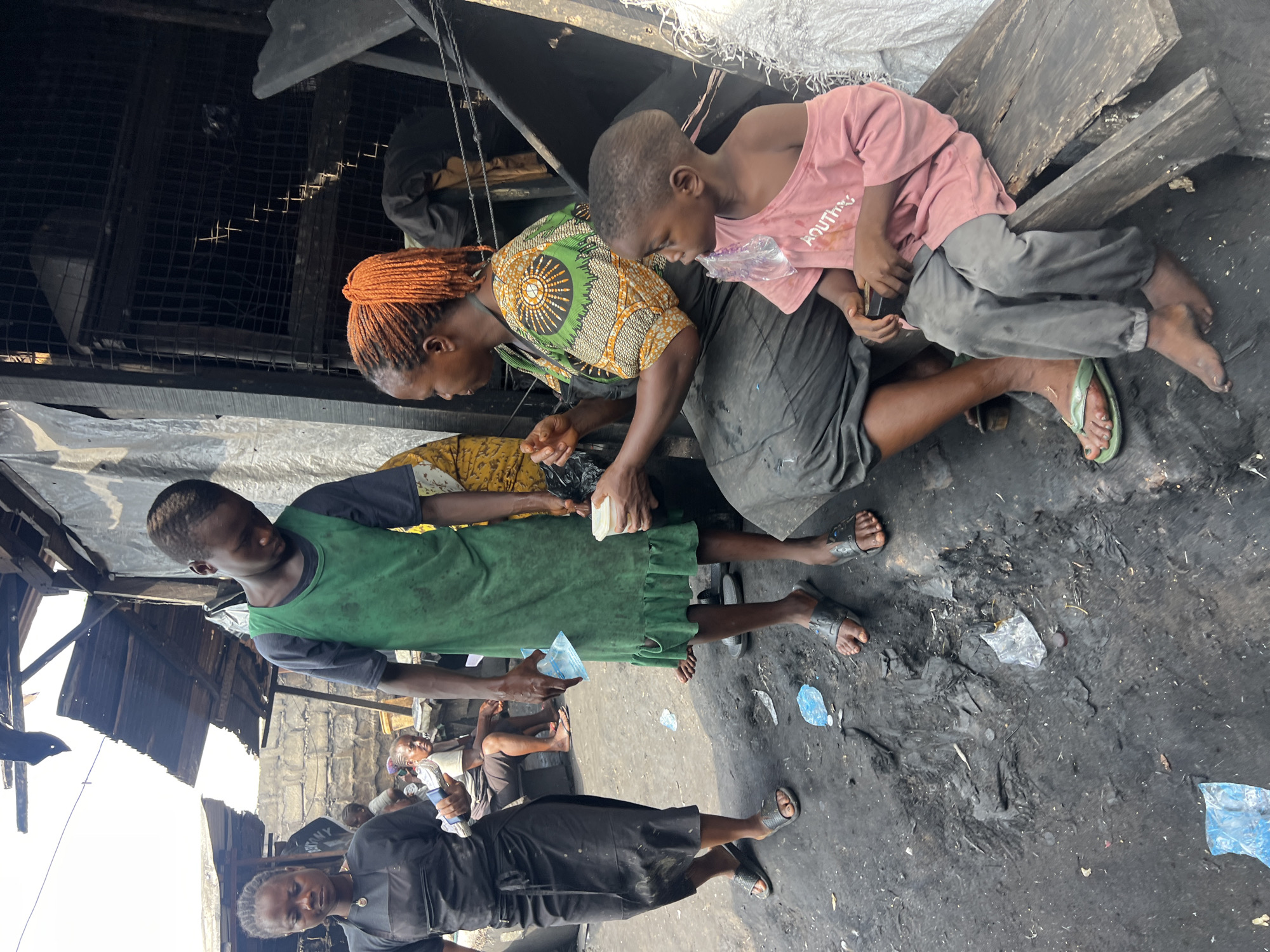 This image captures a scene in Nkpor, Nigeria, likely in a market or an informal settlement, characterized by a rough, dirty, and unpaved ground surface littered with debris and plastic waste. The environment appears somewhat dilapidated, with corrugated metal roofing and wire mesh structures visible in the background, suggesting a functional but basic setting.
The primary focus is on a group of individuals. In the foreground, a young child wearing a pink t-shirt is seated on a rough wooden bench. Next to them, an adult woman with bright orange braided hair is also seated and appears to be interacting with the child, possibly offering food or drink from a small white cup she is holding. The woman is dressed in a patterned top and a dark skirt.
Standing to the left of the seated pair is a young person wearing a green tank top and a dark skirt. They are holding a clear plastic sachet, likely containing water. Another young person, dressed in a black top and a dark skirt, stands further to the left, also holding a similar plastic sachet. In the background, other individuals are visible, including a woman sitting on a bench and another person partially obscured. The overall impression is of everyday life unfolding in a challenging environment.
