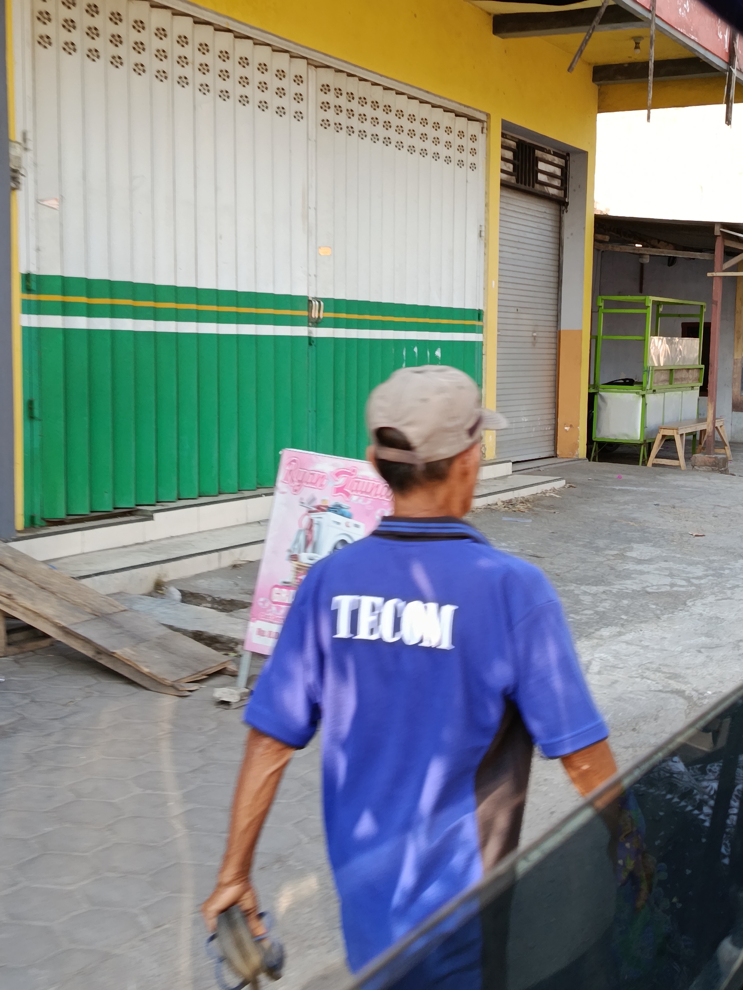 A person wearing a blue t-shirt with the text "TECOM" in white letters on the back and a tan baseball cap is walking away from the camera in the city of Bataan, Indonesia. The person is holding a dark object in their right hand. The background features a yellow building with a white and green striped roll-up door, and a pink sign that appears to be an advertisement for a laundry service. There are also some wooden planks on the ground and a gray, textured walkway.
