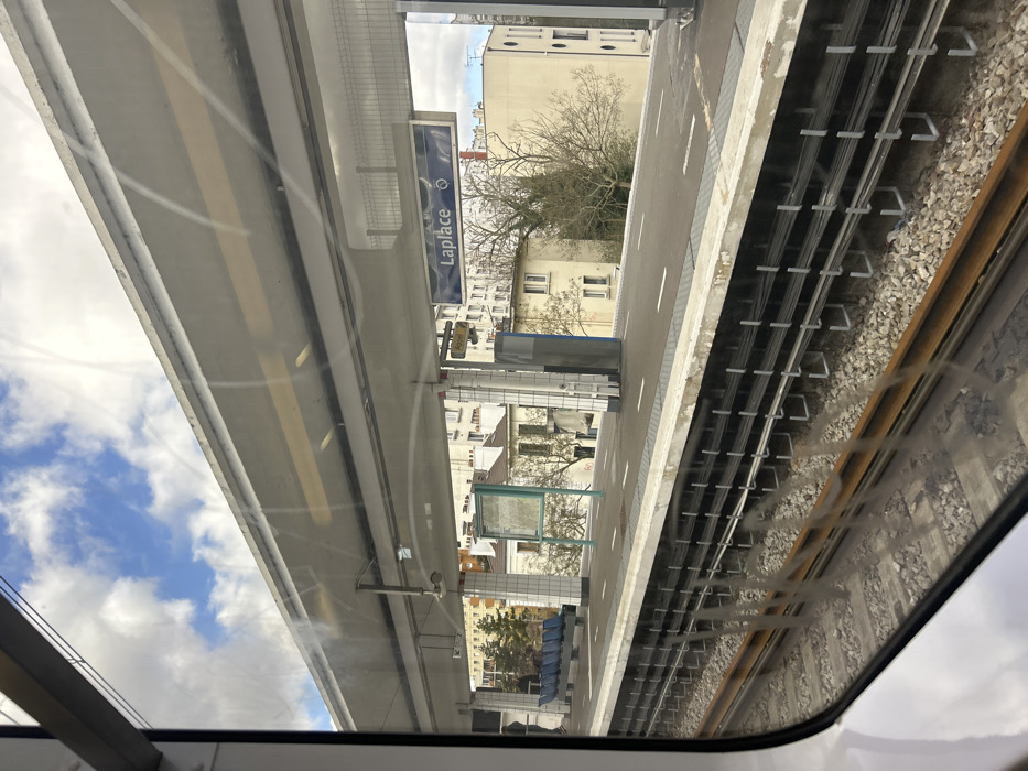 The image displays an elevated train station platform, identified as "Laplace" station in Arcueil, France, viewed from inside a train car through a window with visible smudges and scratches.
The concrete platform runs horizontally across the mid-ground. A blue sign with white lettering clearly displays "Laplace" and a train symbol. An overhead concrete canopy extends across the platform. White square-tiled columns support the canopy. On the left side of the platform, a row of blue benches is visible, with one individual partially discernible near them. A green-framed information board stands on the platform to the right of the benches.
Adjacent to the platform, two parallel train tracks are laid on a bed of gravel ballast. A metallic third rail, or similar conductor rail, runs alongside the tracks. Below the platform edge, a series of cables are secured horizontally along the concrete structure.
In the background, behind the station, multiple light-colored, multi-story residential buildings are visible. Deciduous trees, mostly bare of leaves, and some evergreen foliage are interspersed among the buildings. The sky above is partly cloudy with patches of blue. The scene is illuminated by natural light, indicating daytime.