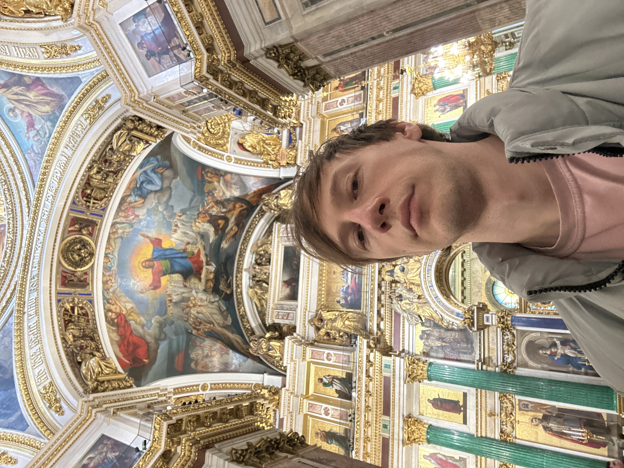 A man is taking a selfie inside the Saint Isaac's Cathedral in Saint Petersburg, Russia. He is wearing a light grey jacket and a pink shirt. The camera is angled upwards, capturing the intricate ceiling details, including paintings, gold decorations, and green columns.