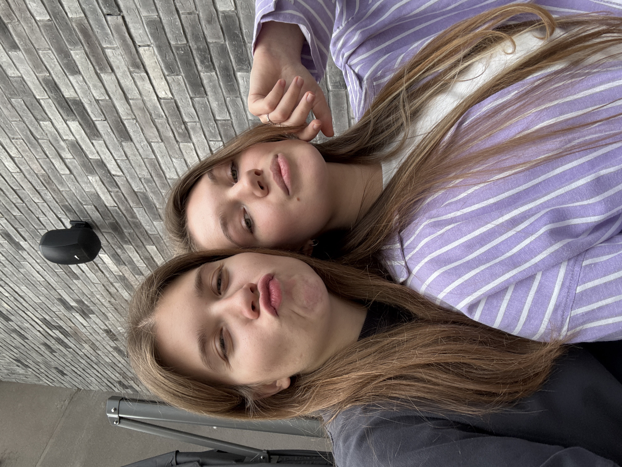 Two young women are posing closely together, likely for a selfie. The woman closer to the camera is pouting her lips, while the other is gazing at the camera with a slight smirk. The top woman is wearing a light purple and white striped collared shirt. The bottom woman is wearing a grey shirt. Their hair is light brown. A portion of a grey brick wall is visible in the background, with a black outdoor speaker mounted on it. Parts of a stroller are visible on the bottom left of the image. Based on the presence of the brick wall and the apparent public setting, this image was likely taken in Belgrade, Serbia.