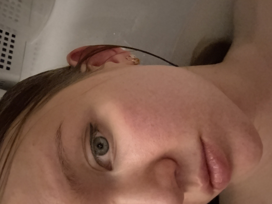 A close-up, angled view of a fair-skinned person's face reveals light gray eyes with dark eyeliner, minimal eyebrow hair, and soft pink lips. The skin appears slightly flushed, with visible pores. A small earring is visible in the upper ear. Brown hair frames the face. The background includes what looks like a white appliance, possibly a washing machine or dryer, with a portion of a white wall or surface also visible. The image is slightly blurry, particularly in the background.