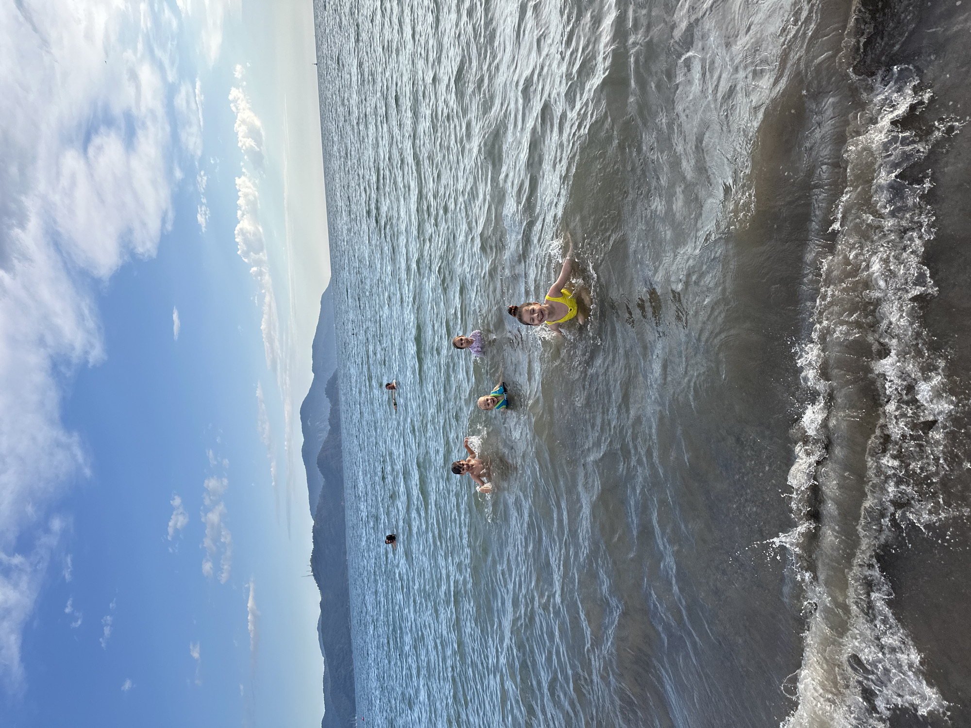 The image depicts a group of people swimming in the sea off the coast of Fethiye, Turkey. Five people are visible in the water at varying distances from the shore. A girl in a yellow swimsuit is closest to the camera and appears to be smiling. Another girl, wearing a white and pink swimsuit, is slightly behind her. A child in a blue swim shirt and goggles is swimming between them. Further back, one person swims with their arms out, and another person is partially submerged. The water is calm but agitated near the shore, showing some wave action. The sky is blue with scattered white clouds. A large mountain range forms the backdrop of the scene. The image appears to be taken during the daytime under bright sunlight.