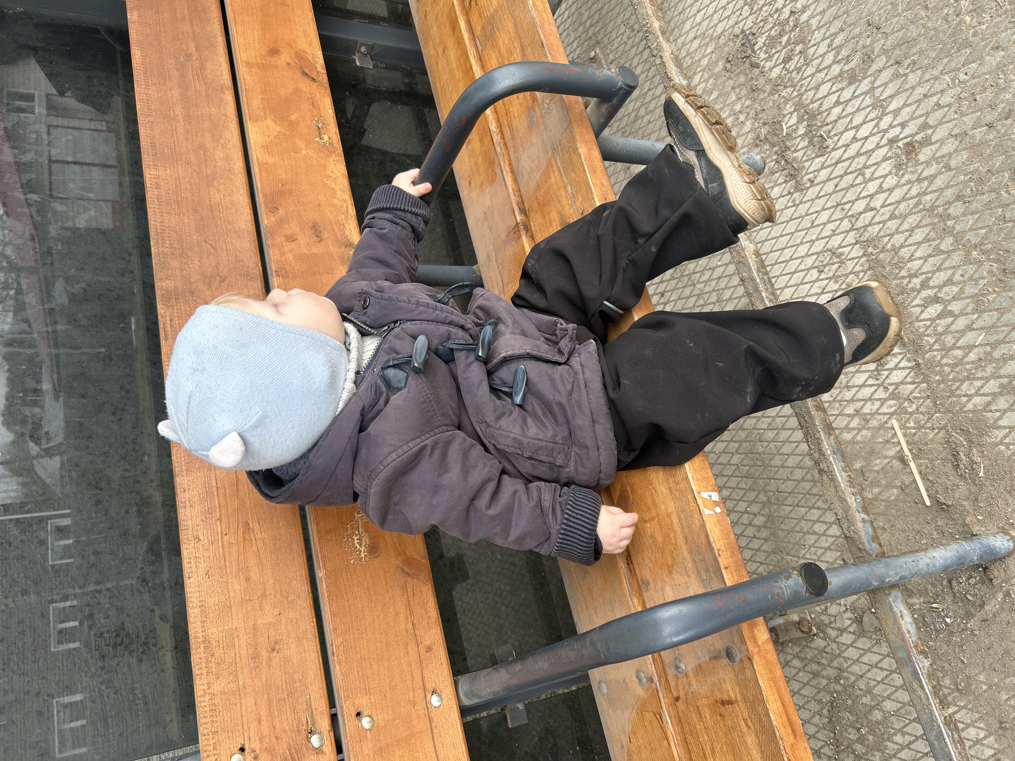 A child is lying on a park bench in Chaikovsky, Russia. The child wears a light blue hat with small ears, a dark gray coat with toggle buttons, black pants, and dirty sneakers. The child's left hand is gripping a curved gray metal support bar. The bench is made of wooden planks, and beneath it is a gray metal grate with accumulated dirt. Part of a building with large windows is visible in the background.