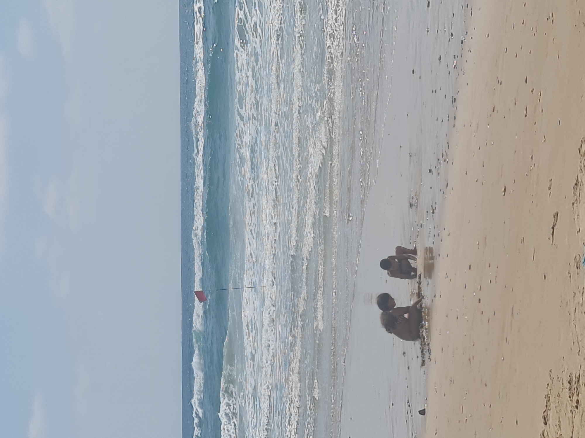 The image captures a serene beach scene with two children playing in the shallow water. The sky is clear and blue, with a few scattered clouds. The ocean is a vibrant turquoise, with gentle waves rolling towards the shore. A red flag is visible in the distance, planted on a pole in the water, possibly indicating a warning or a specific zone. The sand on the beach is a light beige color, scattered with small shells and pebbles. The children are crouched down, engrossed in their play, their backs to the camera. The overall atmosphere is one of peacefulness and childhood innocence.
