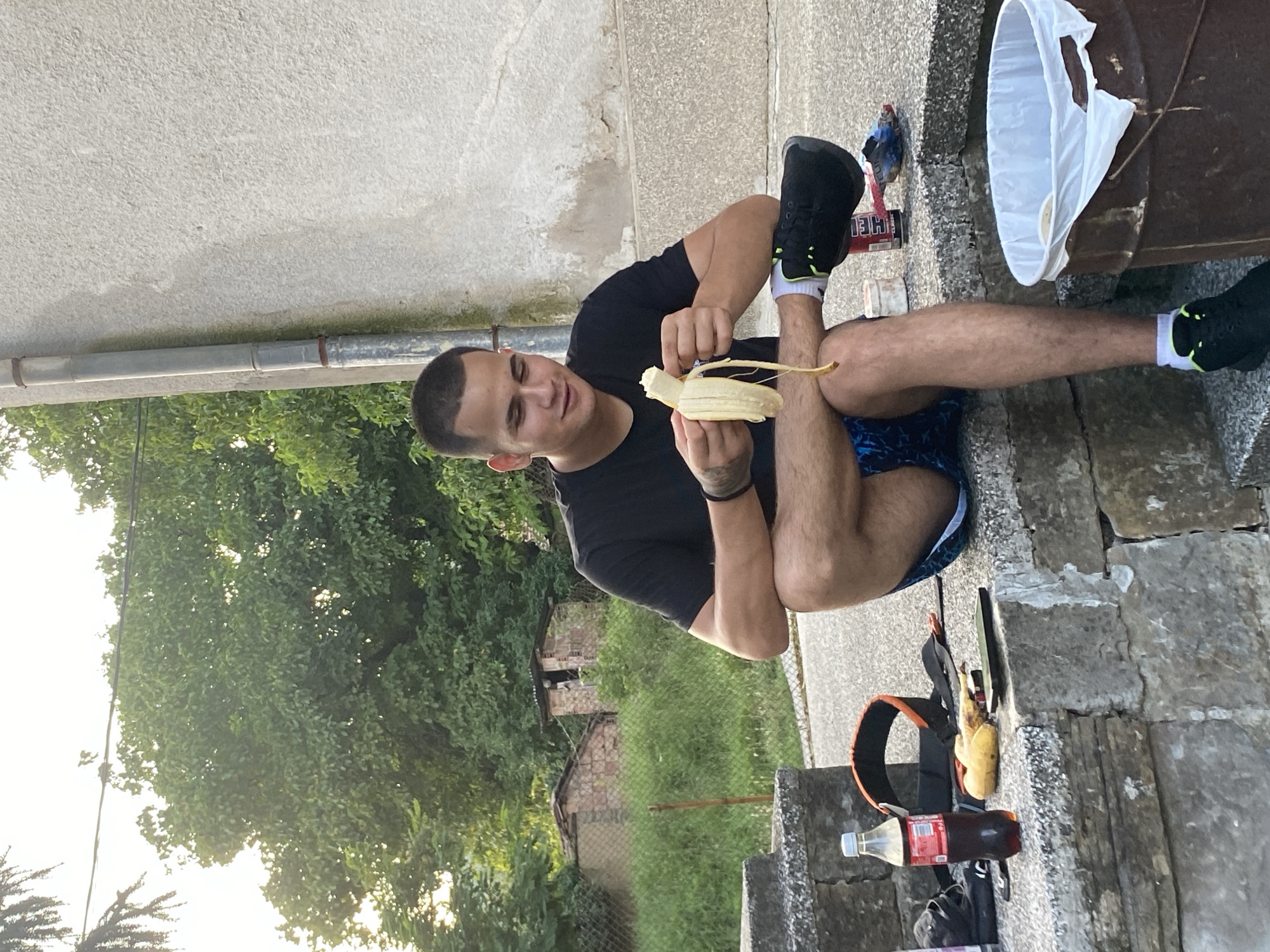 In Hirevo, Bulgaria, a young man with a close-cropped haircut is seated on a stone step, peeling a banana. He is wearing a black t-shirt and blue patterned shorts. His legs are crossed, and he is looking down at the banana in his hands. In front of him, on the step, is a bottle of Coca-Cola, a banana, and a phone. Behind him, a wall of a building and lush green trees are visible. A trash can with a white liner is to his right. The setting appears to be outdoors, possibly a residential area.