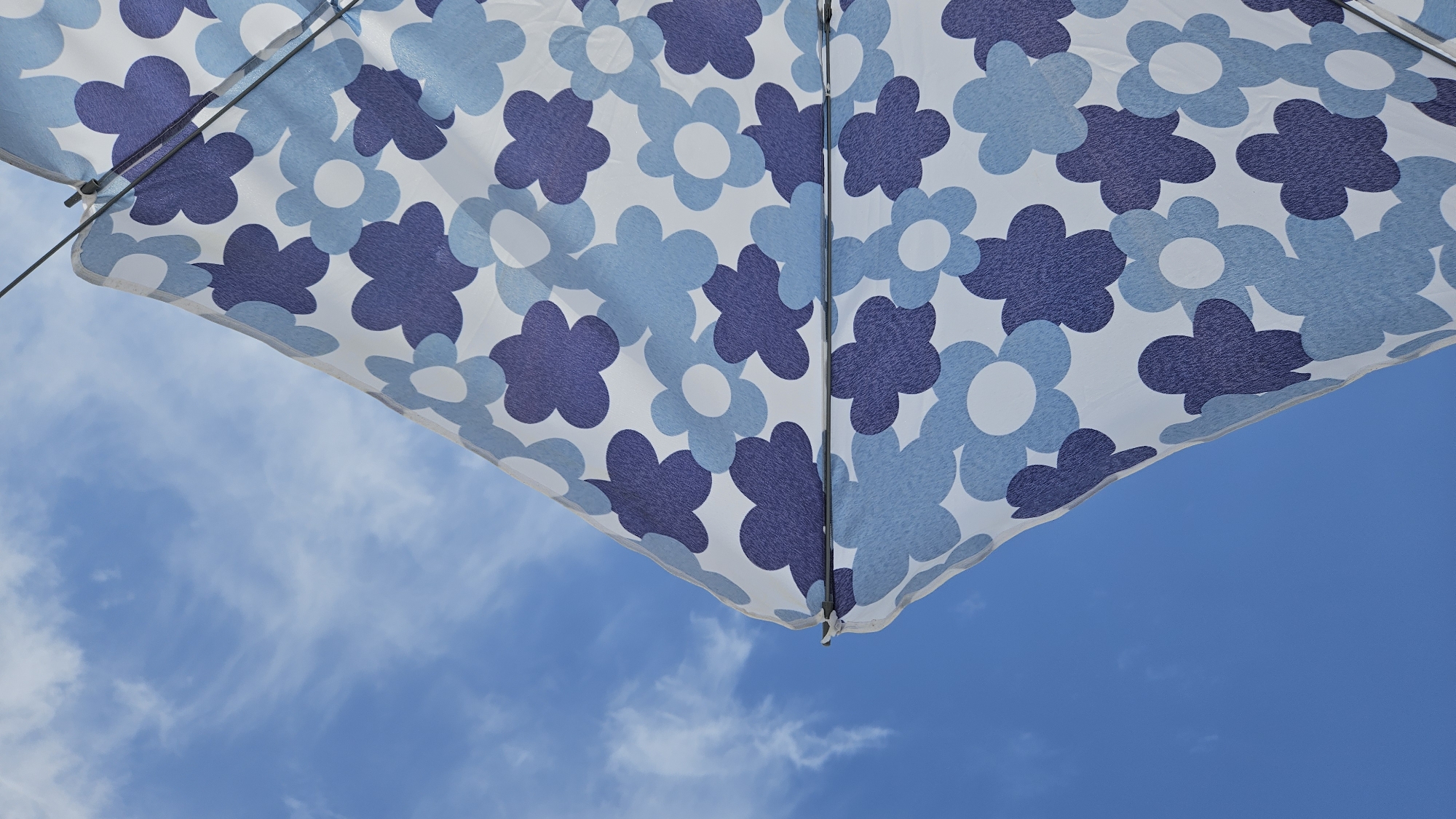 An extreme close-up, angled shot shows the underside of a white umbrella with a blue floral pattern against a bright blue sky filled with wispy white clouds. The umbrella is partially open and angled upwards, with its fabric creating a large triangular shape that dominates the right side of the frame. The floral pattern consists of stylized, four-petaled flowers in varying shades of blue, including a deep navy and a lighter sky blue, scattered across the white fabric. The metallic ribs of the umbrella are visible in the upper right corner. The sky takes up the left portion of the image and is a clear, vibrant blue, streaked with thin, white clouds that create a sense of movement. No people or identifiable landmarks are present. The image appears to have been taken outdoors in a location with bright daylight, possibly L'Île-Rousse, France, given the clear sky and outdoor setting often associated with coastal towns.