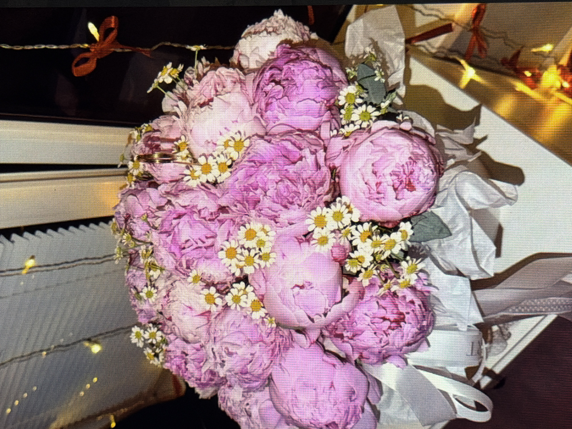 The image depicts a close-up, vertical view of a vibrant bouquet of pink peonies interspersed with small white daisies. The flowers are arranged in a white decorative box, adorned with white and grey ribbons. In the background, a gray, ridged surface with faint bokeh lights is visible. To the right, a dark background features a red ribbon tied to a thin string, with warm, out-of-focus lights illuminating the scene. The location is not discernible from the image.