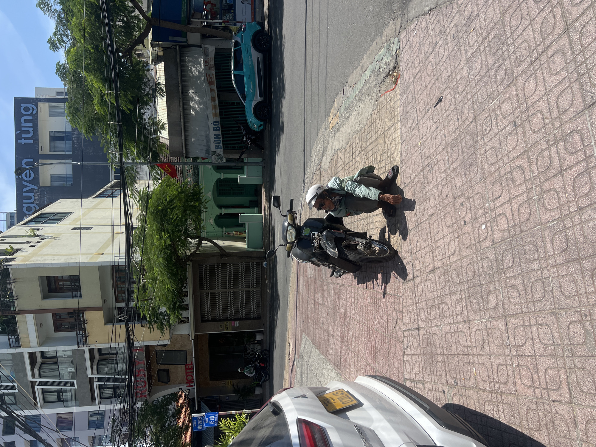A man wearing a white helmet, light green jacket, and dark pants is crouched next to a black motorcycle on a red-tiled sidewalk. The sidewalk runs alongside a street with a parked light blue car. Behind the motorcycle, buildings are visible, including a storefront with the words "BUN BO." Across the street, a multi-story building is seen with a black sign. The scene appears to be outdoors on a sunny day. A white car is partially visible on the bottom right of the frame. The street name "LE LOI" is seen on a blue sign attached to a building. The location is the Nha Trang Ward in Vietnam.