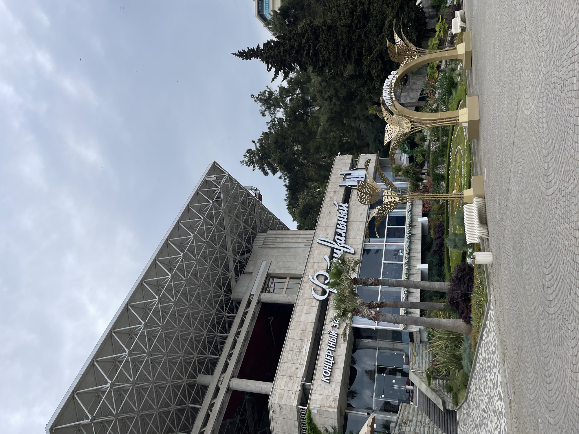 The image shows the Primorsky Concert Hall in Sochi, Russia, taken from a low-angle perspective. The building features a modern architectural design with a triangular, lattice-like roof structure. A sign bearing the concert hall's name in Cyrillic script adorns the building's facade. In front of the building is a decorative golden arch structure with plant decorations and manicured greenery. A paved walkway is visible in the foreground. The sky is overcast. There is no apparent sign of people.