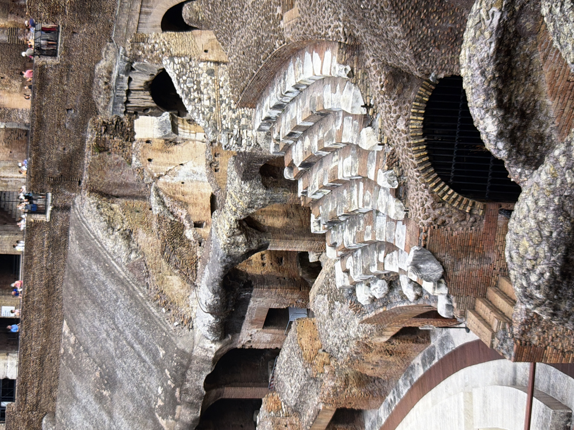 The image depicts the interior of a large, ancient stone amphitheater, likely the Colosseum in Rome. The architecture features weathered brickwork, arched openings, and tiered seating constructed from stone and brick. Some sections of the seating are intact, showing rows of steps and resting places. Other areas show signs of decay and damage, with crumbling stones and exposed structures. In the background, a railing is visible with several individuals standing and observing the scene. The overall impression is one of historical grandeur and significant age. The image does not show any elements related to Kyiv, Ukraine.