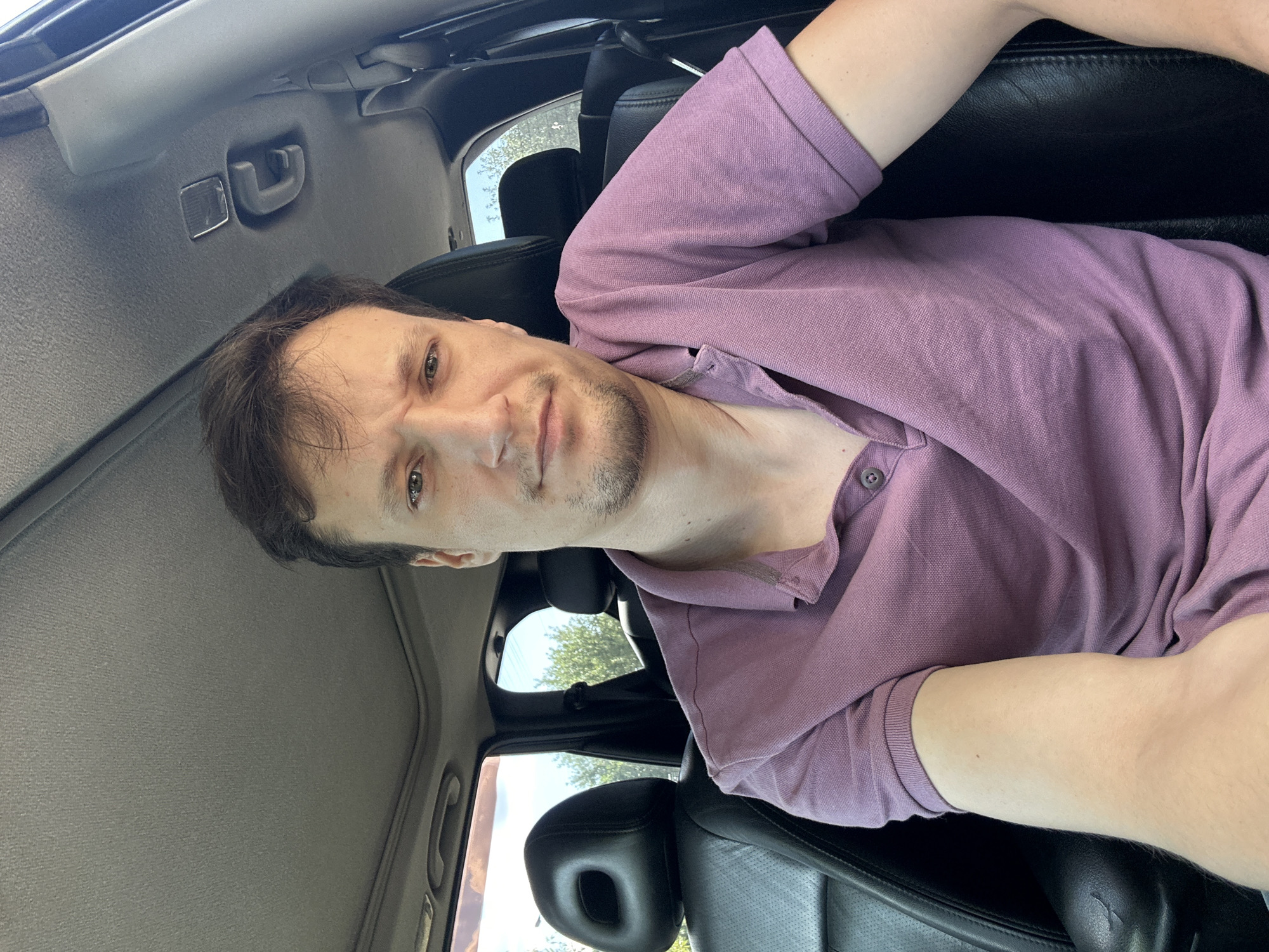 A man in a purple polo shirt is sitting in the driver's seat of a car. The interior of the car is visible, including black leather seats and the sunroof. The man is looking at the camera with a neutral expression. Outside the car, trees and sky are visible through the windows. The image appears to be taken during the daytime. There is no indication of the specific location in Makiv, Ukraine.