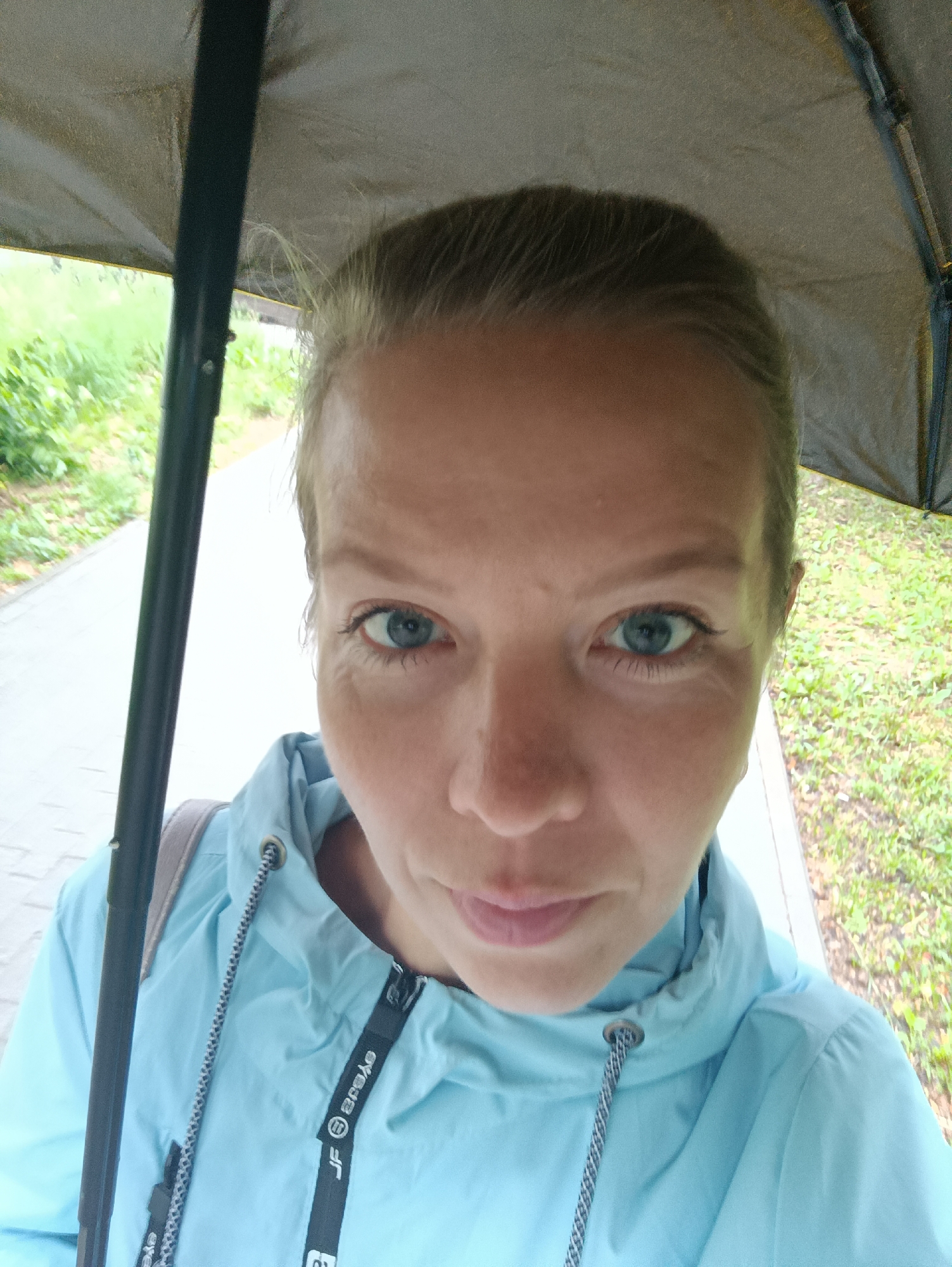 A woman takes a close-up selfie outdoors in Chaikovsky, Russia, holding a dark-colored umbrella over her head. She looks directly at the camera with a neutral expression, her blue eyes prominent. Her hair, which appears light blonde or light brown, is pulled back from her face. She is wearing a light blue hooded jacket, possibly a windbreaker, with black and grey drawstrings around the hood. On a black fabric strap running along the zipper of her jacket, the text "JF Byebye" is visible, with a reversed "ǝʌɐǝq" also present. A backpack strap is visible over her left shoulder. The background features a paved path on the left and green grass or foliage on the right, suggesting a park or outdoor recreational area. The presence of the umbrella implies overcast or rainy weather during the daytime.