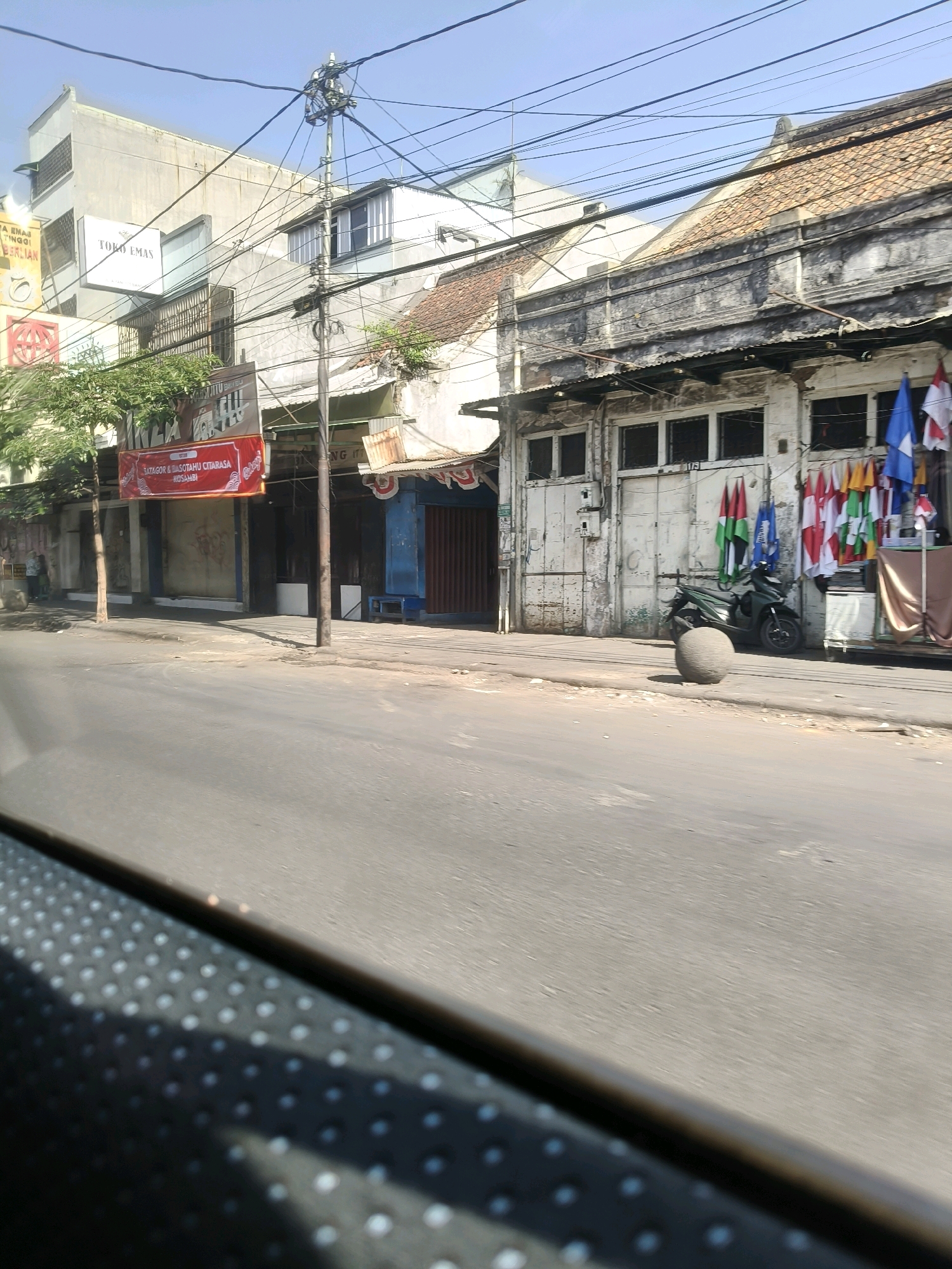 The image depicts a street scene in Bandung City, Indonesia, likely in an older commercial area. The foreground is dominated by the blurry interior of a vehicle, with a textured surface of raised dots visible. The middle ground shows a paved road and sidewalk. On the right side of the sidewalk, there is a green motorcycle parked, and in front of it, a large, round, grey stone object. The buildings lining the street appear to be old, with some showing signs of wear and tear. Flags, including Indonesian flags and possibly others, are displayed outside one of the buildings. Signs are visible on some of the buildings, including one that reads "TOKO EMAS" (Gold Shop) and another red banner advertising "BATAGOR & SOTAHU CITARASA KOSAMBI." Trees with green foliage are present along the sidewalk, and power lines crisscross the sky. The sky is clear and blue, indicating a sunny day.
