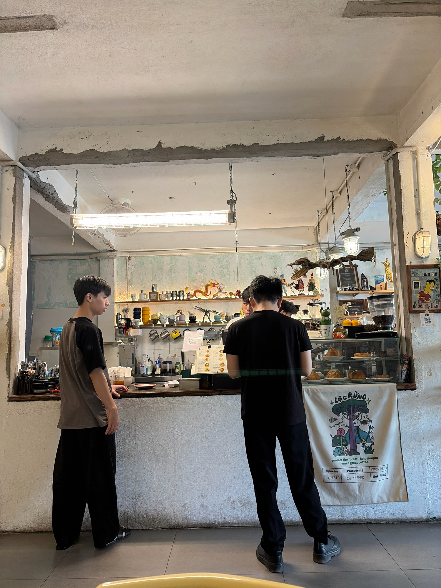 This image depicts the interior of a cafe, likely in Da Nang, Vietnam. Two young men are present. One, wearing a grey and black t-shirt and black pants, stands at the counter looking to the left. The other, in a black t-shirt and black pants, stands at the counter with his back to the camera, facing the display case. The counter features a glass display case filled with pastries, a coffee machine, and various beverage preparation items. Behind the counter, shelves are stocked with mugs, decorative items including a dragon figurine, and other assorted objects. A poster with a nature-themed illustration and text, including "protect the forest - help people make great coffee" and details about coffee processing, hangs on the wall. The cafe has an industrial aesthetic with exposed concrete structures, white-painted walls, and overhead fluorescent lighting.
