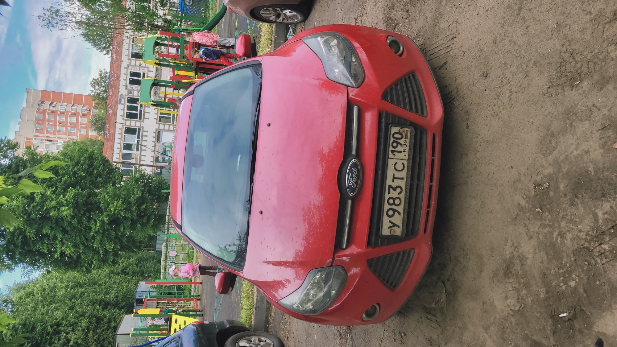 A red Ford car is parked on a dirt lot in front of a playground. The playground has brightly colored structures and is surrounded by trees. In the background, there are apartment buildings. Several children are visible on the playground and near the car. The car's license plate is partially obscured. The sky is overcast.