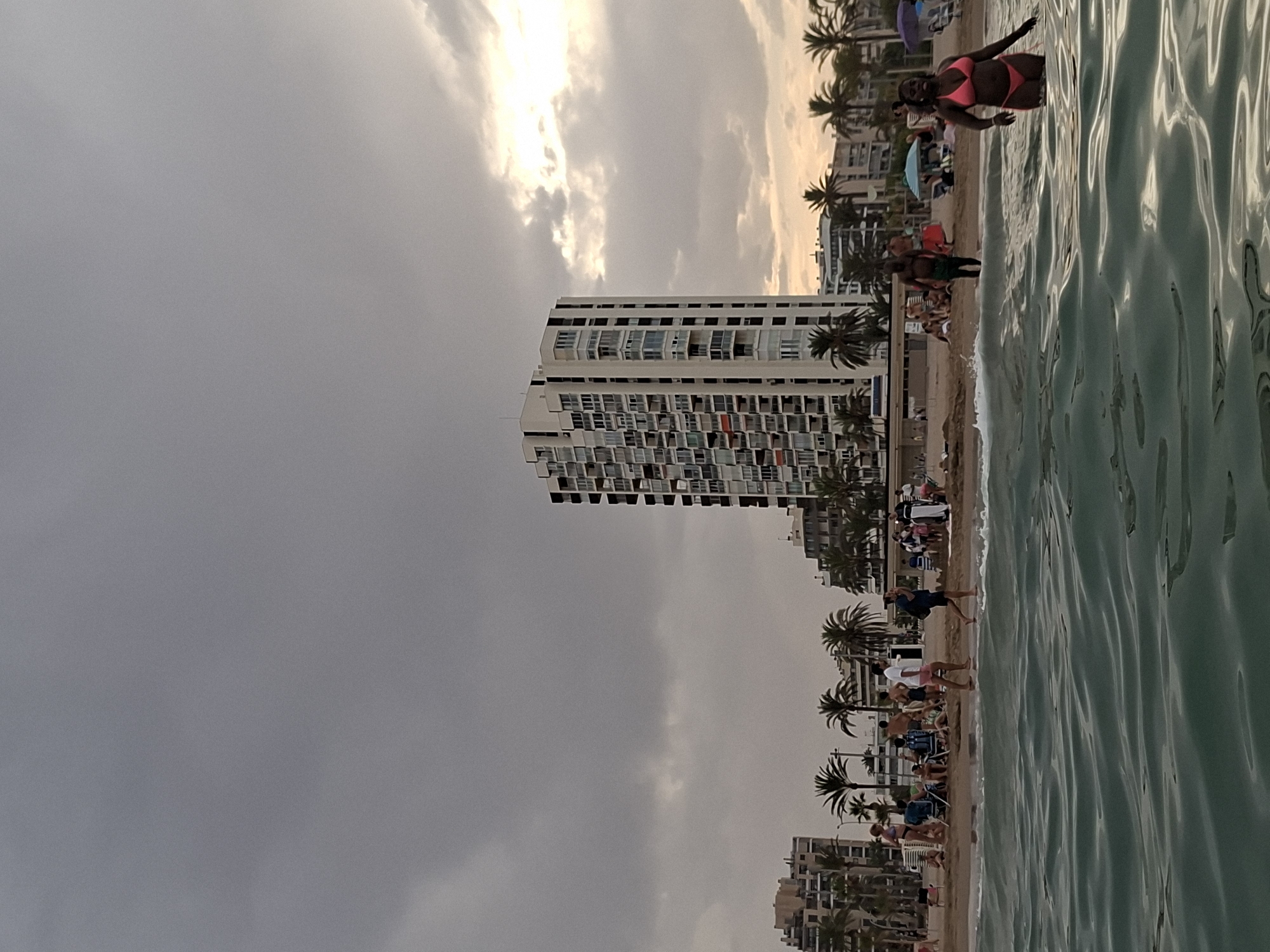 The image depicts a beach scene with a woman in a pink bikini wading in the water. The water is a dark green and appears to be choppy. In the background, there are several buildings, including a tall white high-rise building with balconies. Palm trees are scattered throughout the scene. There are people on the beach, some sitting in chairs and others walking. The sky is cloudy with a patch of bright light peeking through on the right side. The scene is likely a coastal area in the city of Alicante, Spain, possibly near a promenade or beachfront development.